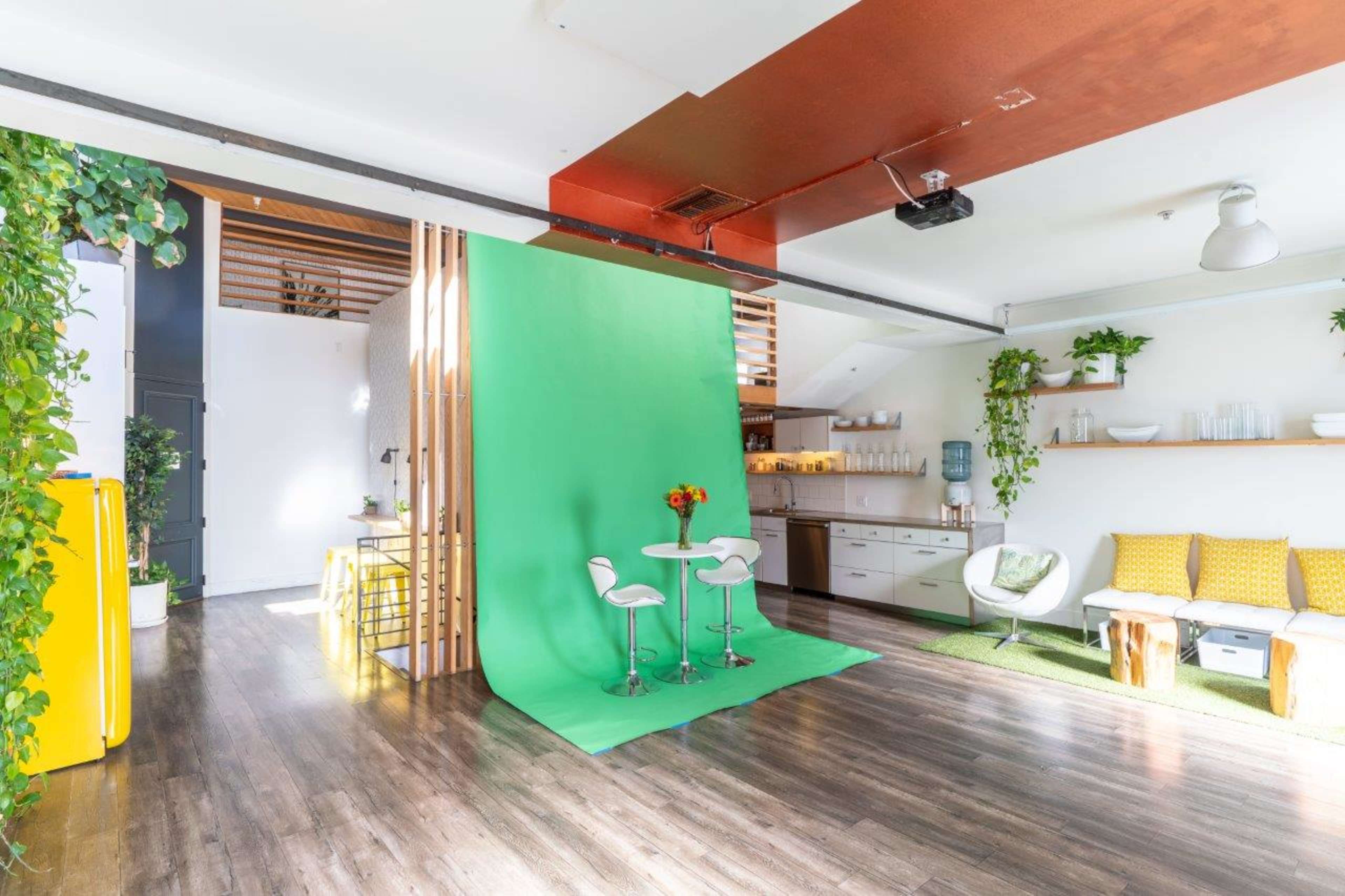 A bright, modern studio space with a green backdrop, two white stools, and a kitchen area, surrounded by plants and decorated with yellow pillows.