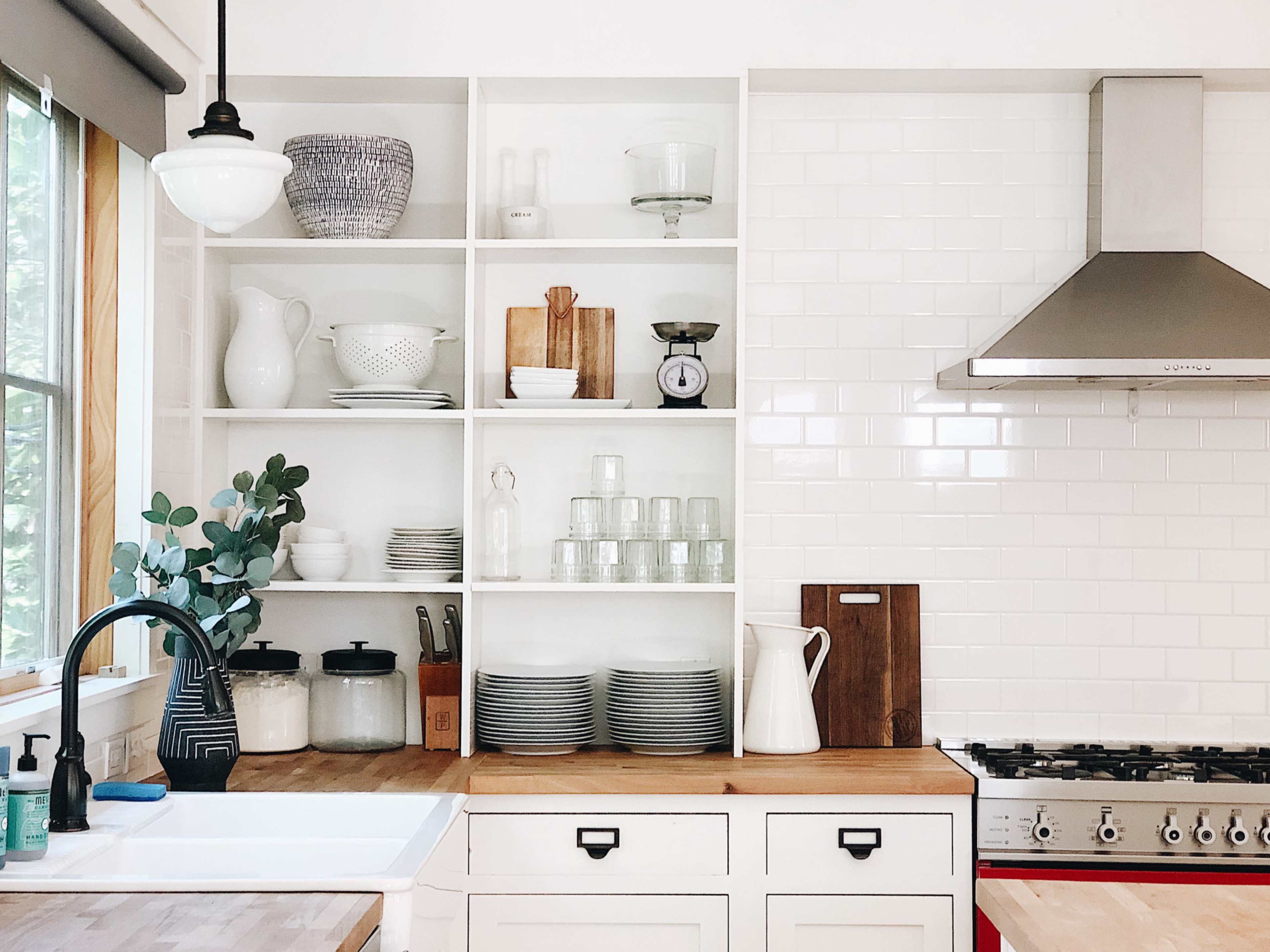 A modern kitchen with open shelving displaying various dishware, glassware, and kitchen tools above a wooden countertop and a stainless steel stove.