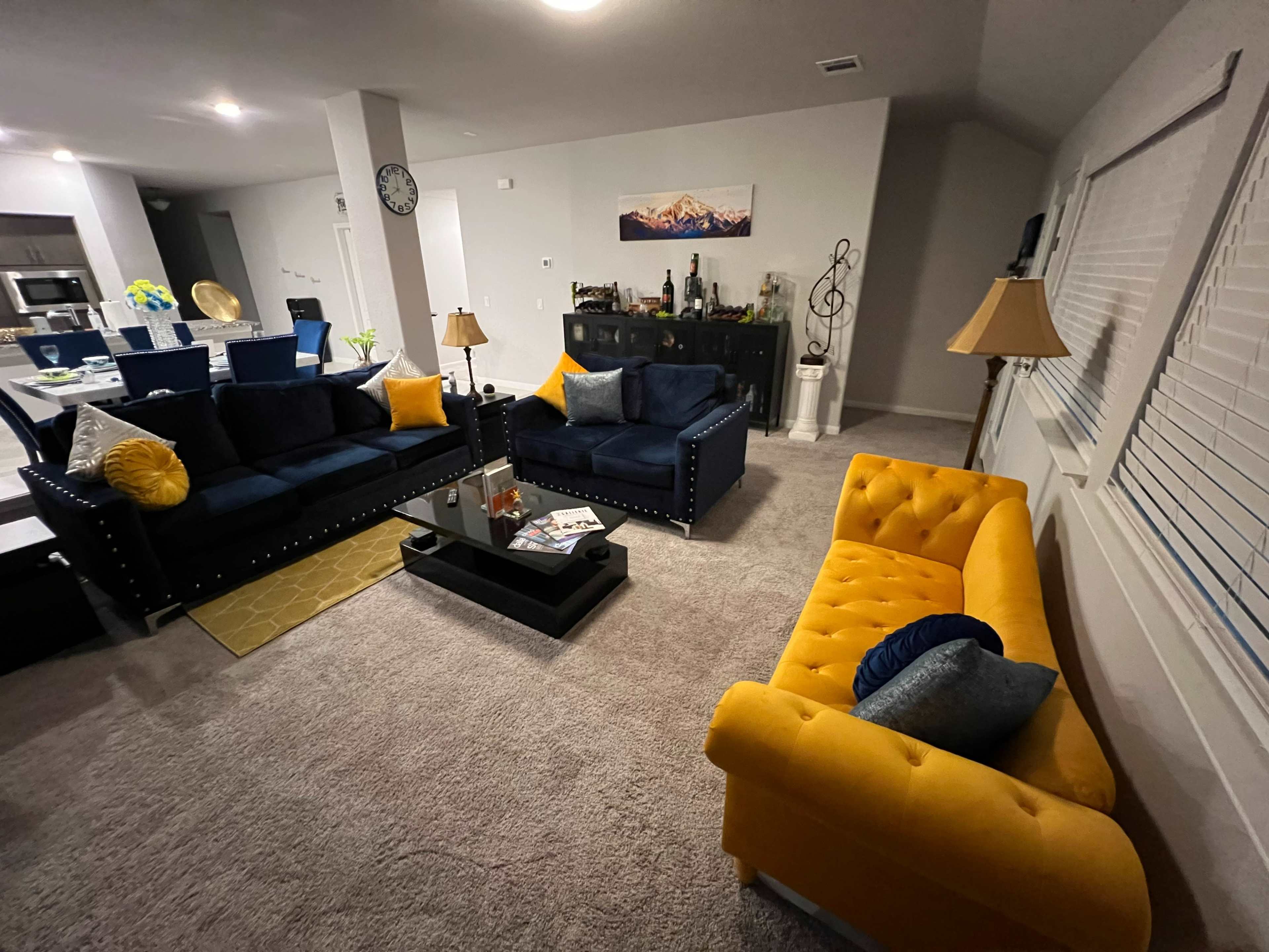 A living room with two navy blue sofas and a yellow loveseat arranged around a coffee table, with a dining area visible in the background.