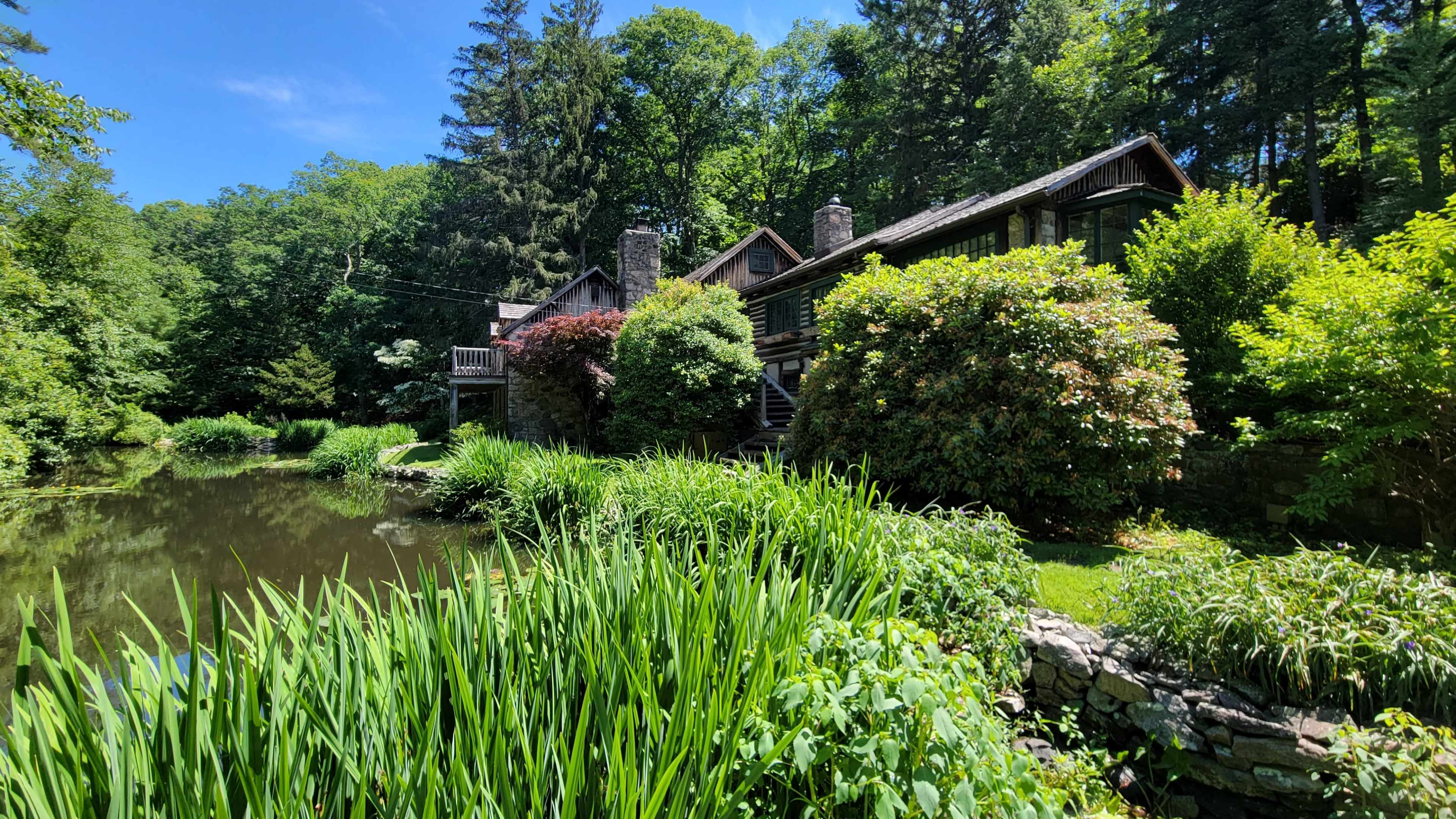 A wooden cabin and a two-story house are surrounded by lush greenery and a pond in a forested area.