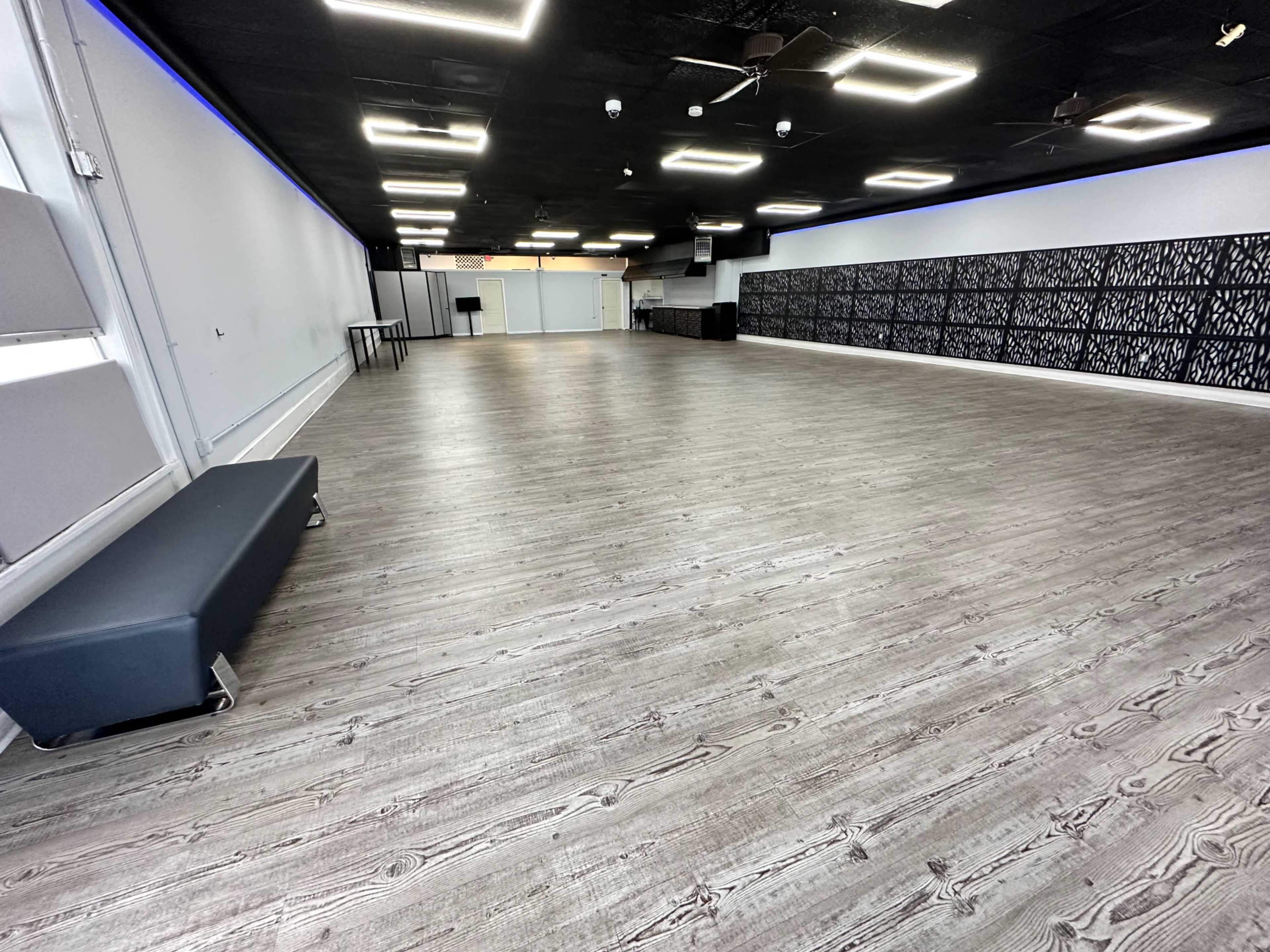 A spacious, empty room with wooden flooring and modern lighting, featuring gray walls and a textured panel along one side.
