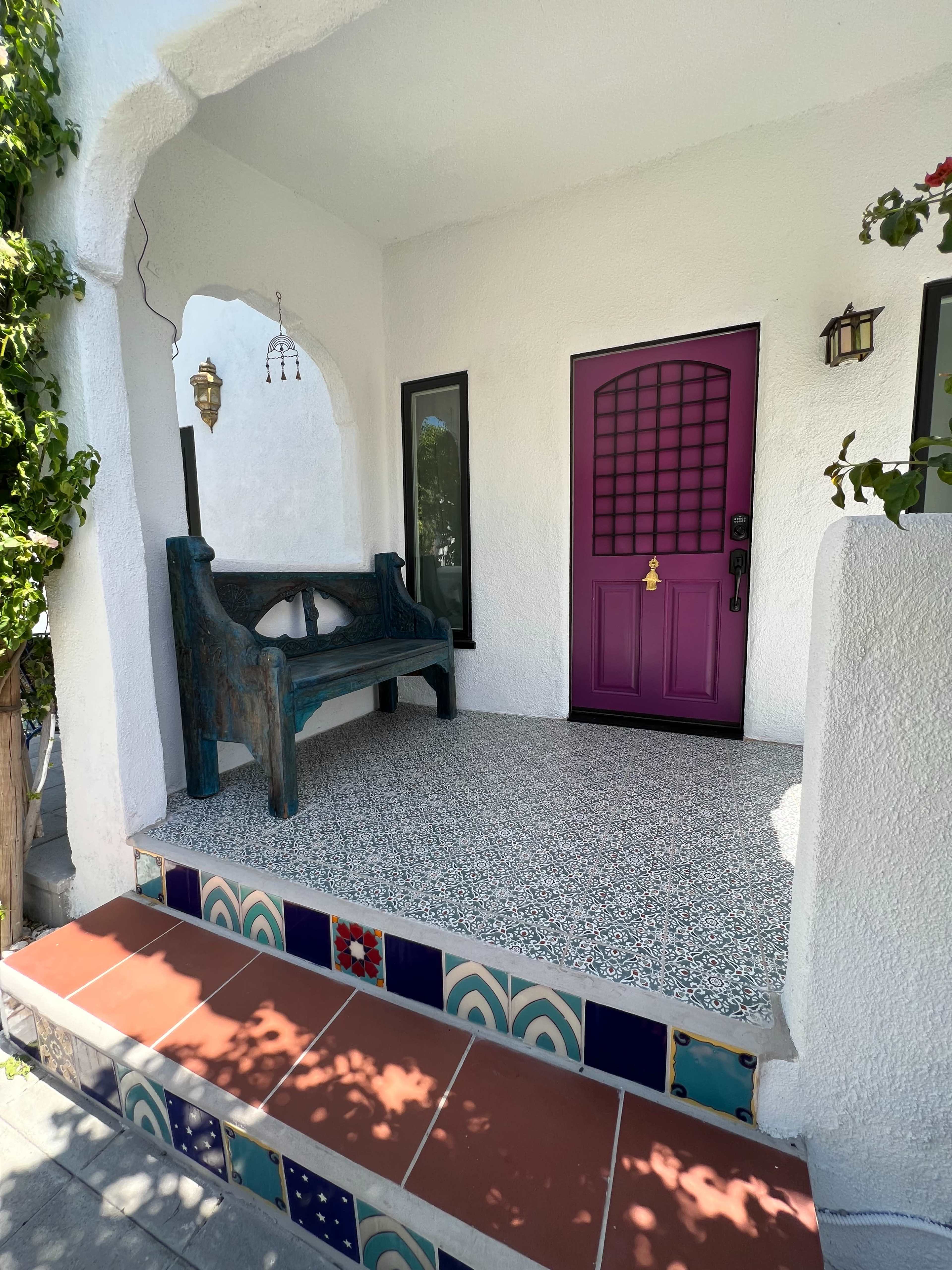 A porch with a purple door, a rustic blue bench, and tiled steps featuring colorful designs.