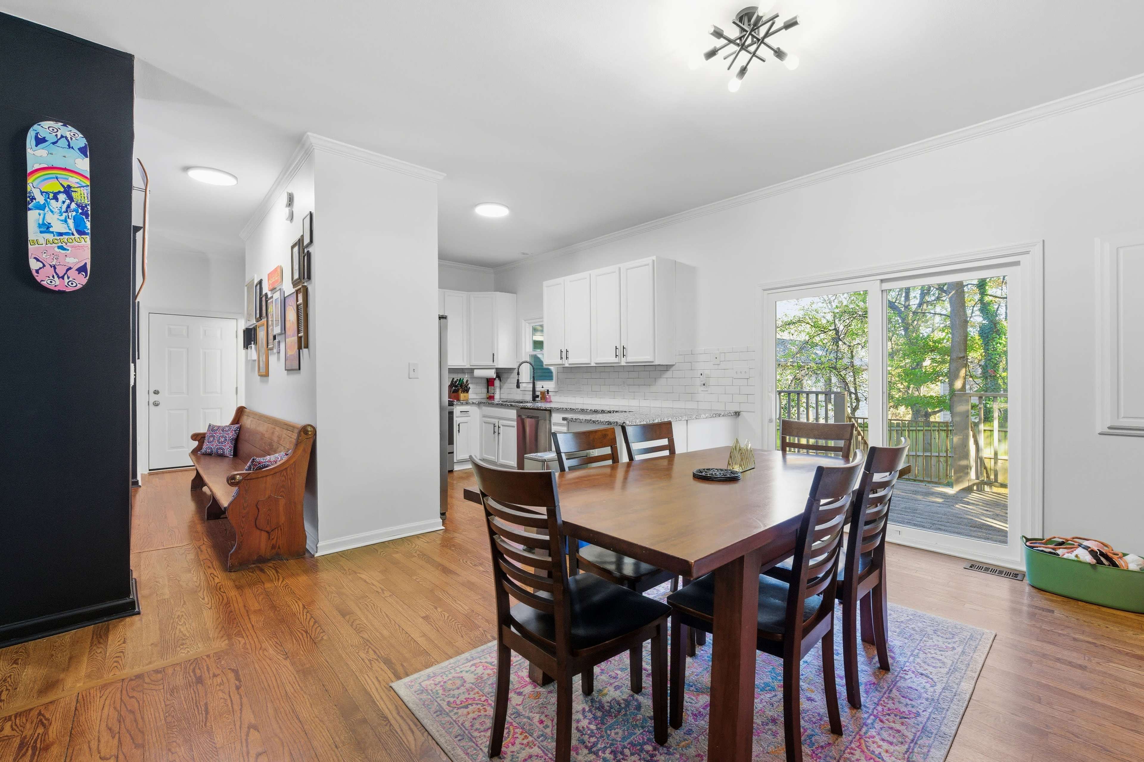The image shows a bright dining area with a wooden table and chairs, leading to a kitchen and an outdoor patio through sliding glass doors.