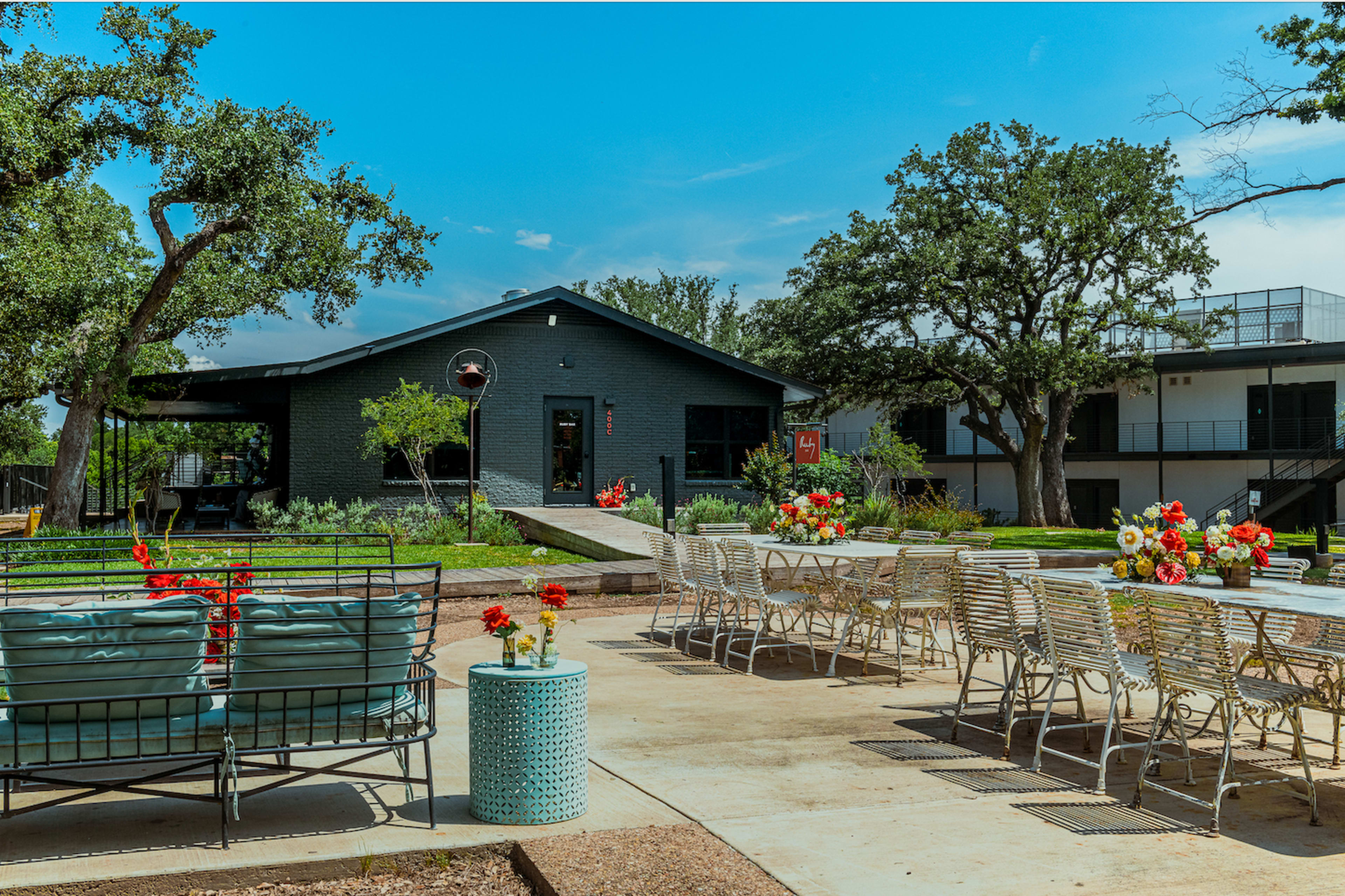 The image shows a modern outdoor space featuring a black building surrounded by greenery, equipped with tables and chairs adorned with colorful flowers.