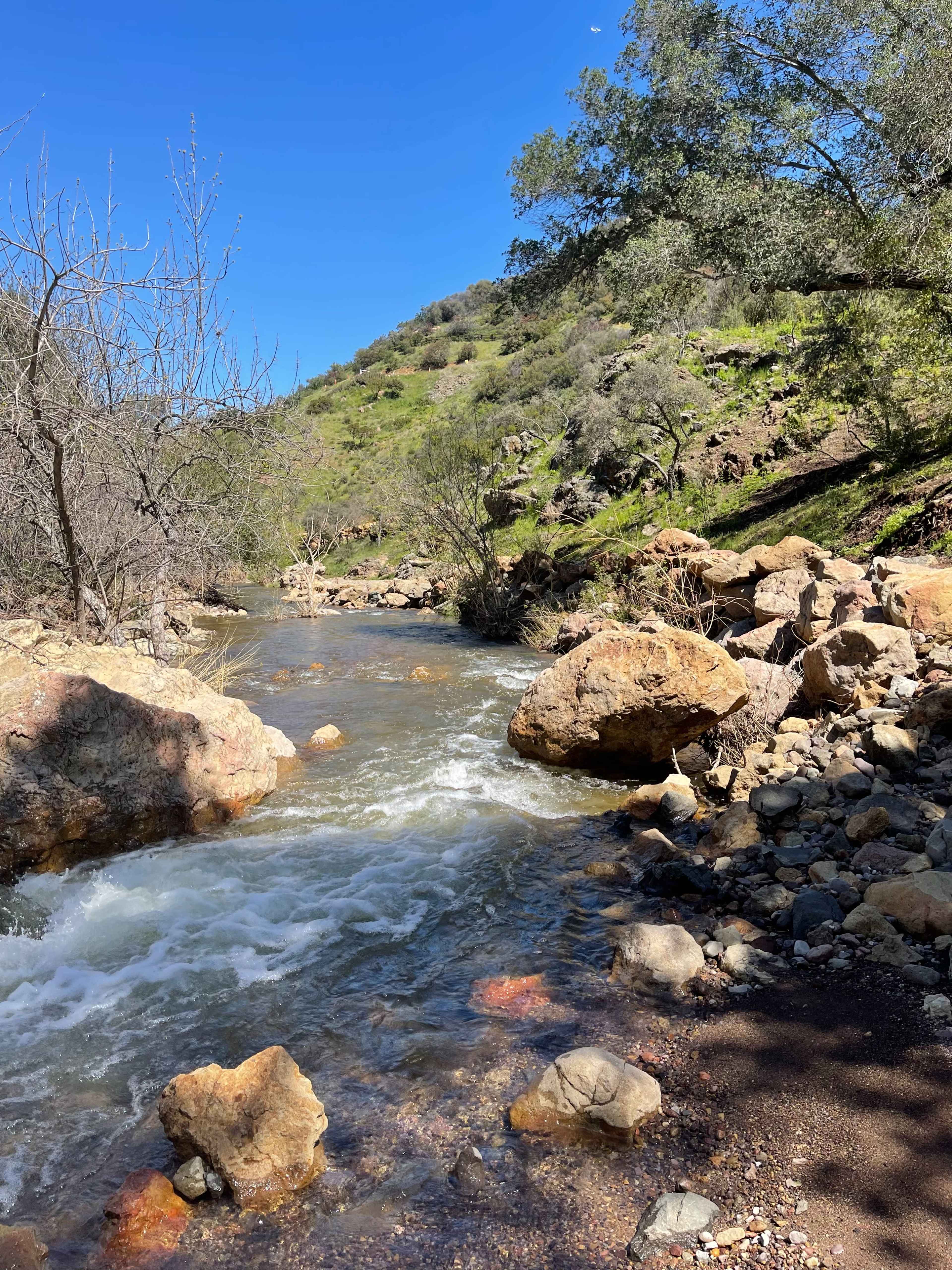 A flowing river surrounded by rocky banks and hillside vegetation under a clear blue sky.
