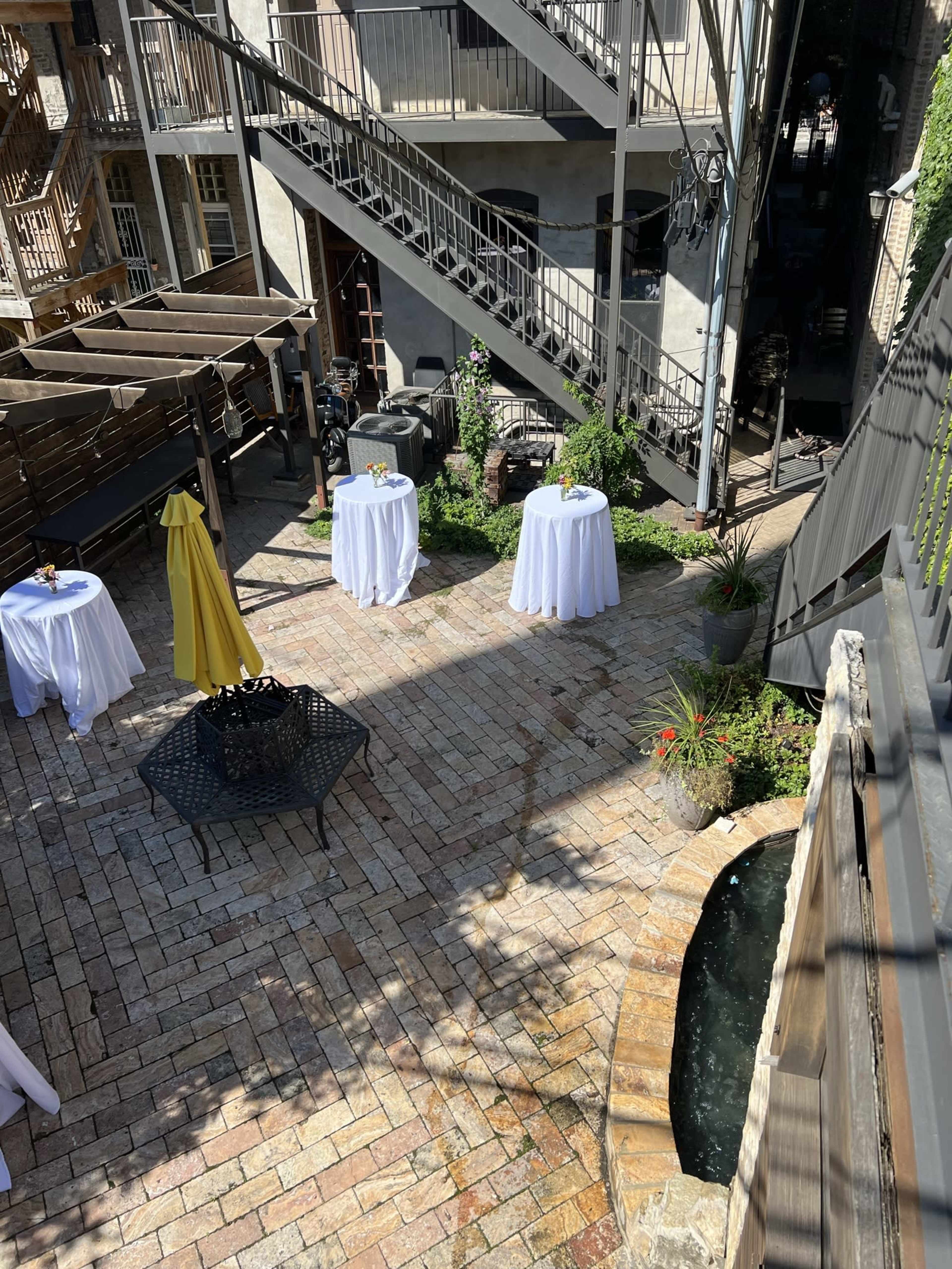 The image shows a courtyard with several round tables dressed in white tablecloths, surrounded by greenery and featuring a small water feature along the brick pathway.