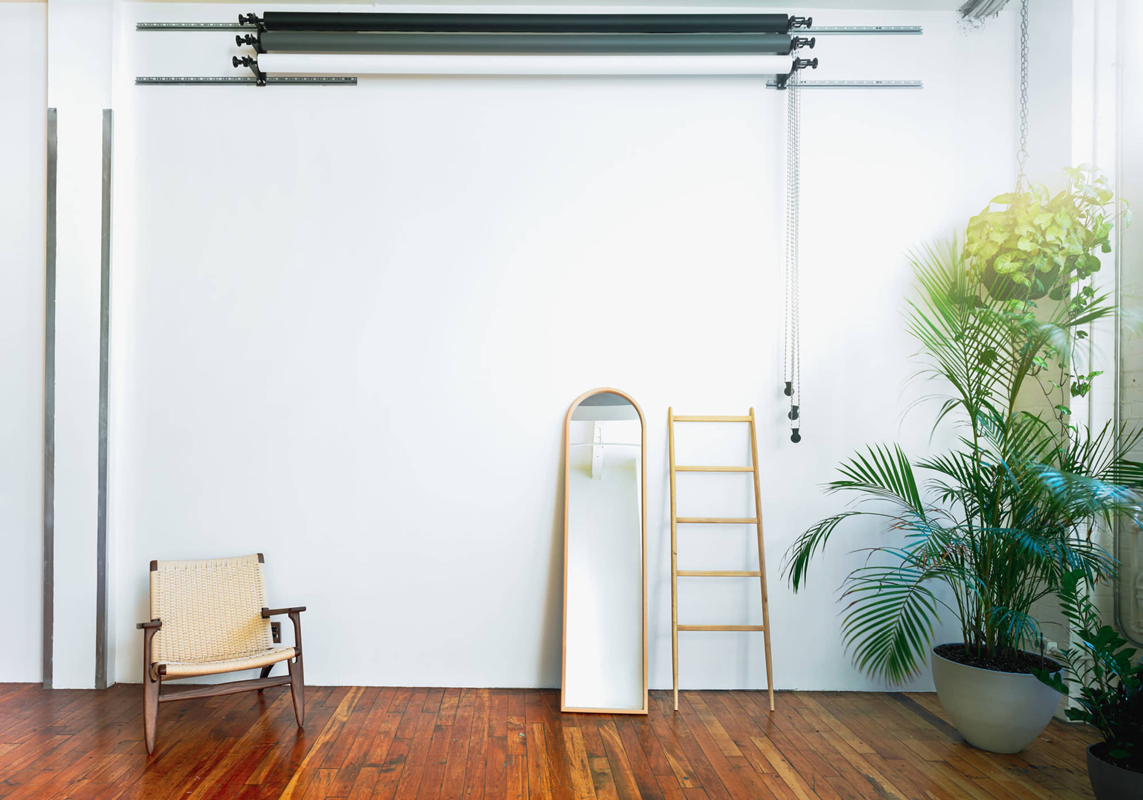 A minimalist interior with a wooden chair, a tall mirror leaning against a wall, a ladder, and a potted plant next to a large window.