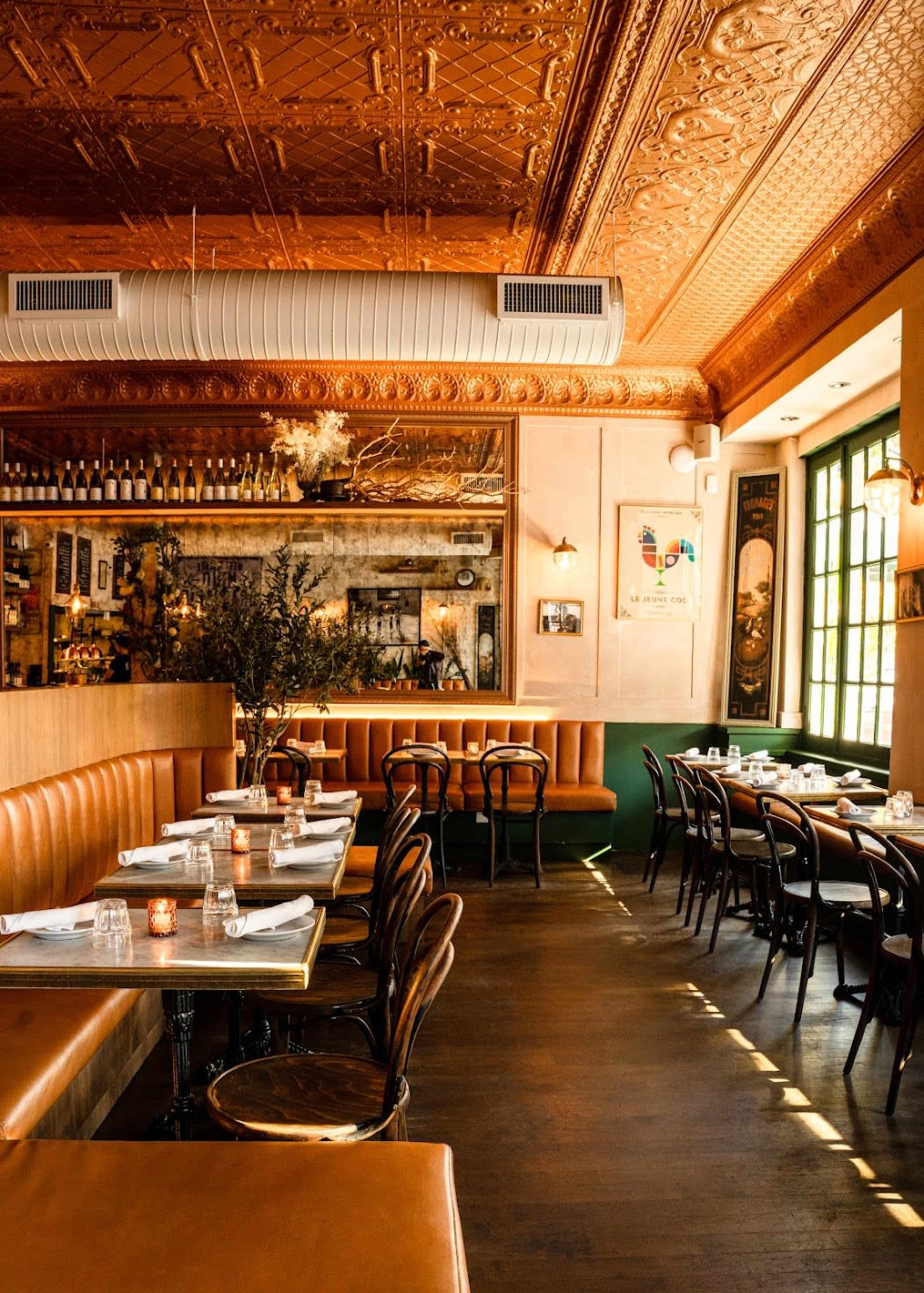 The interior of a restaurant features leather benches, wooden tables, and ornate ceiling details, with soft lighting and a bar in the background.
