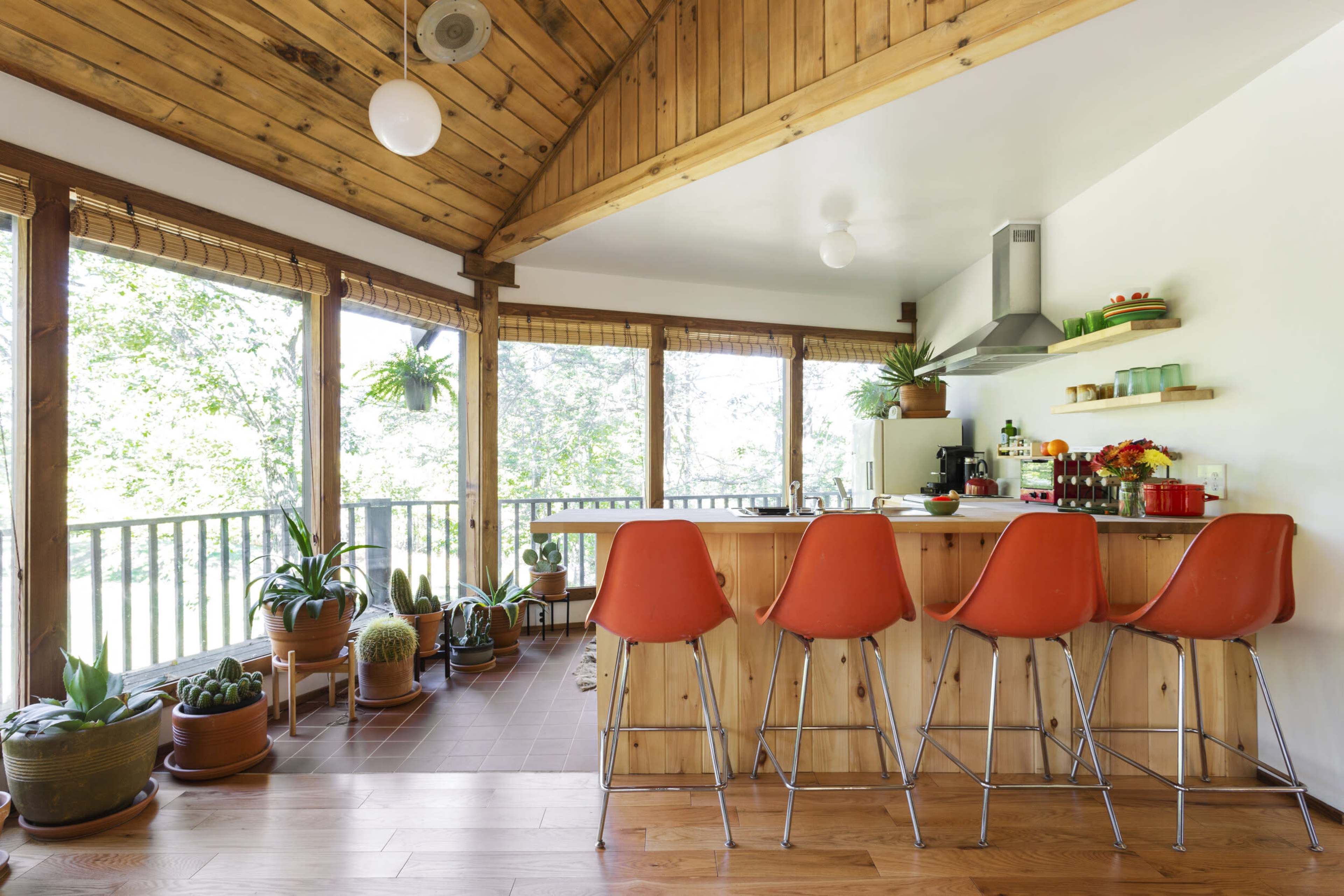 A modern kitchen with a wooden ceiling, featuring an island bar with four orange stools and large windows surrounded by potted plants.