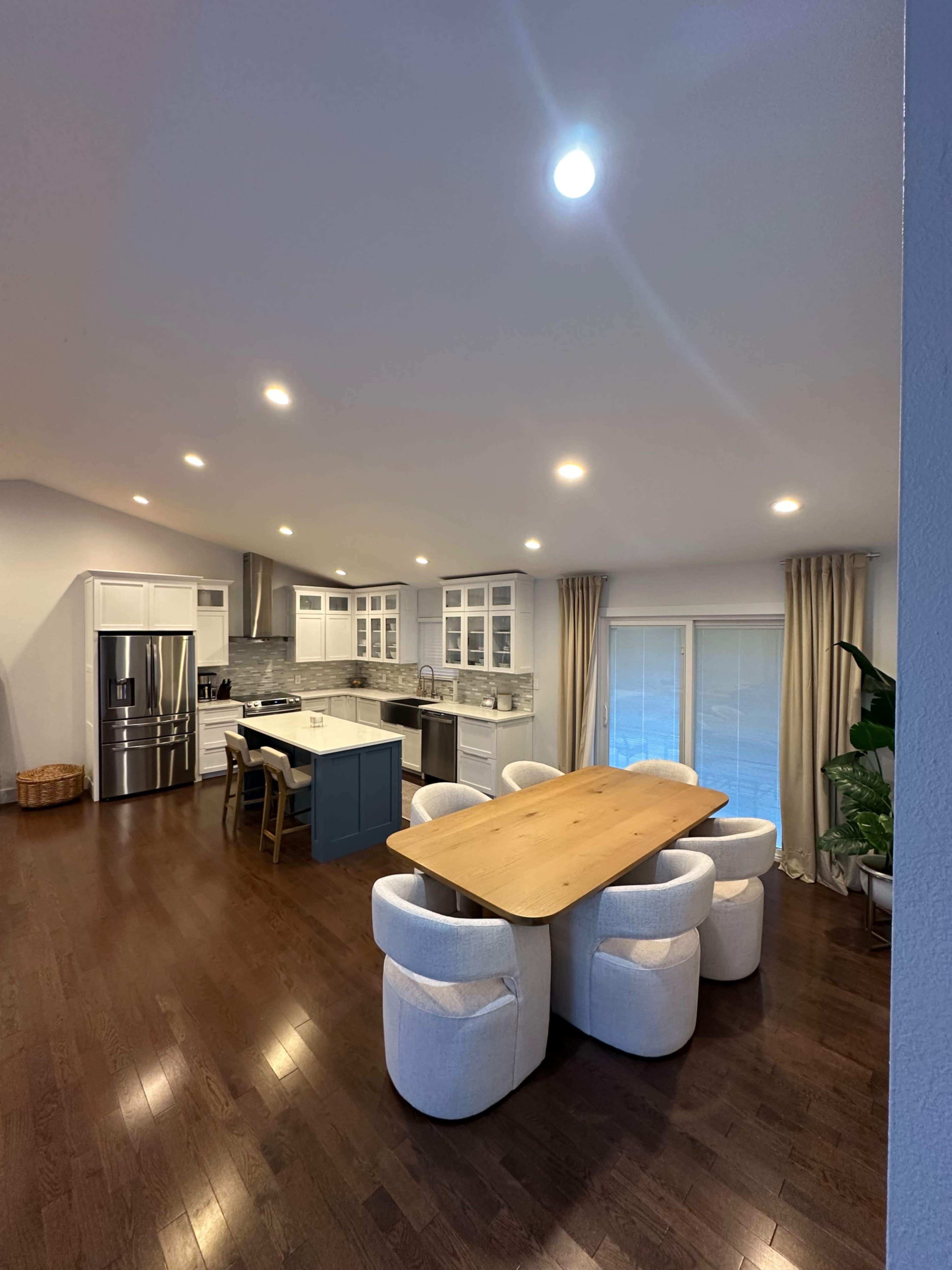 A modern kitchen features a large wooden dining table surrounded by curved chairs, with stainless steel appliances and white cabinetry in the background.