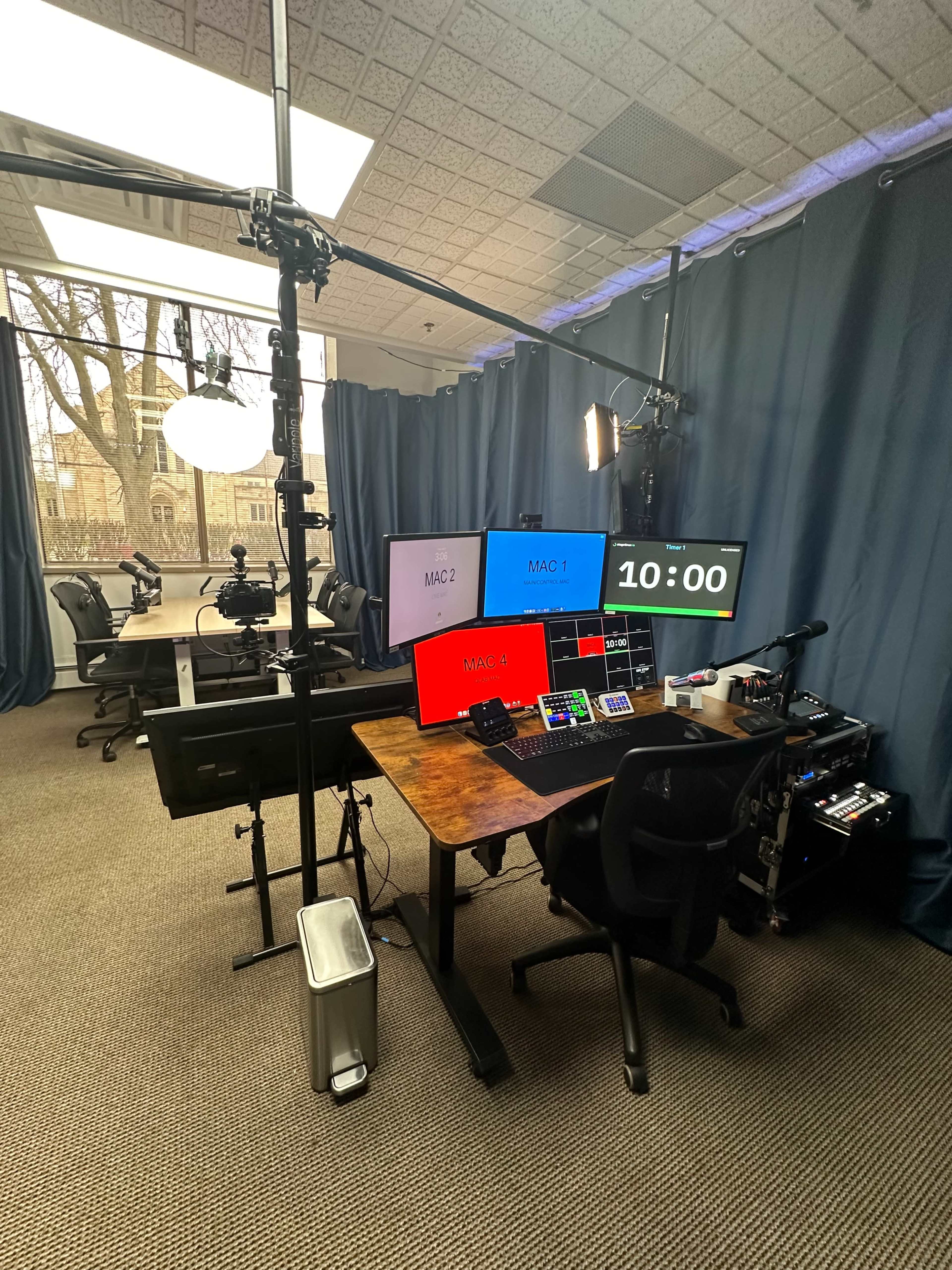 A multimedia production studio equipped with multiple screens, a control panel, and camera setups, surrounded by dark curtains in a room with large windows.