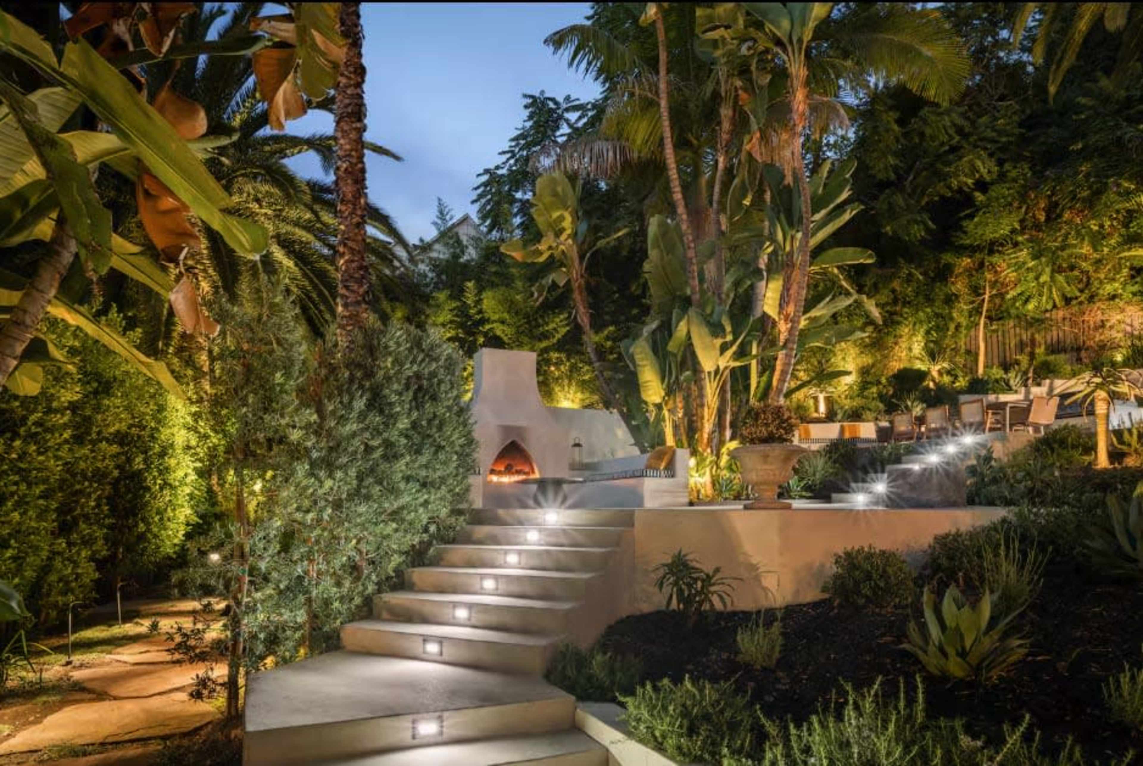A landscaped garden features stone steps illuminated by lights, leading to a seating area surrounded by tropical plants and trees.
