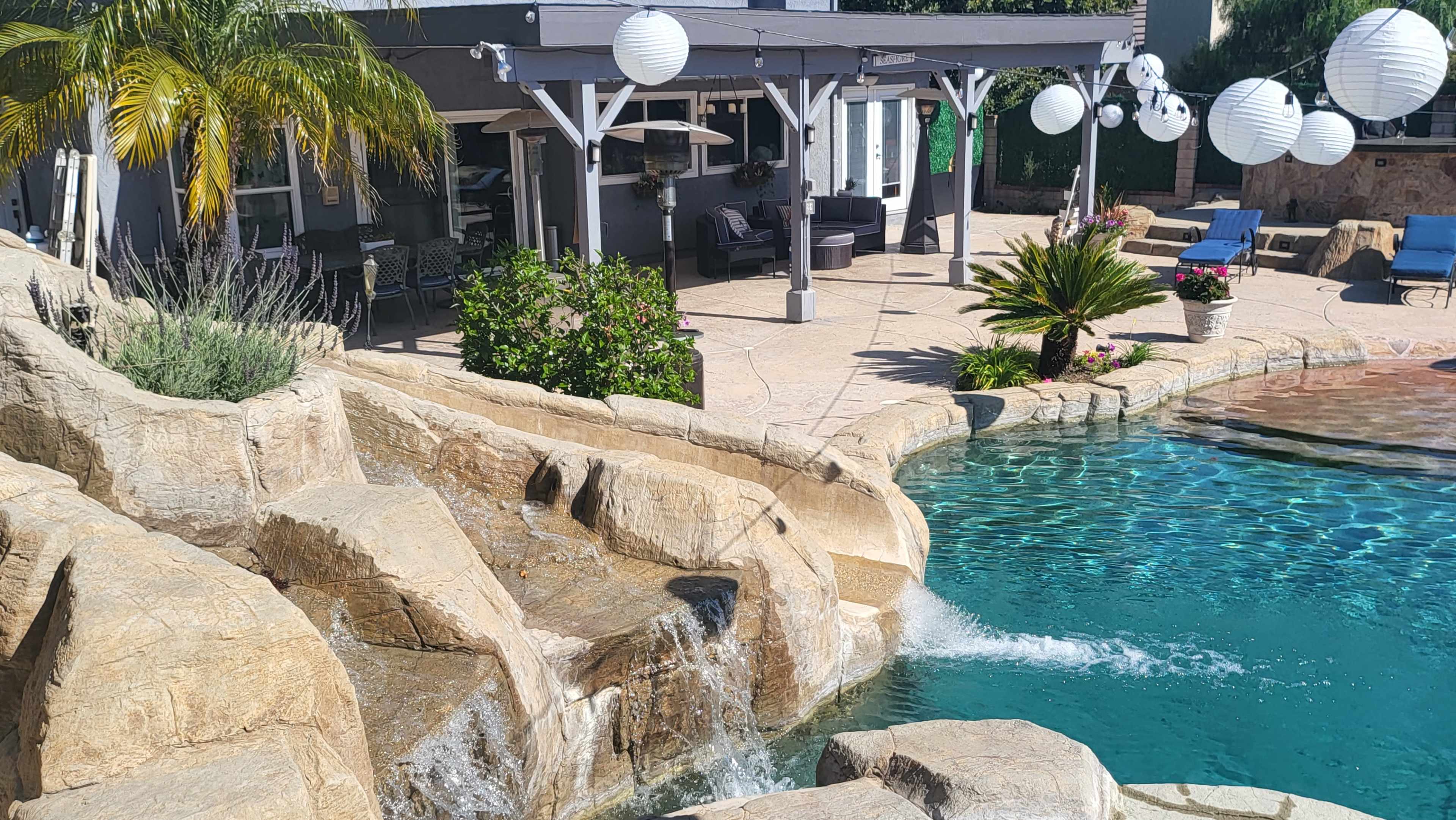 A stone waterfall flows into a turquoise swimming pool surrounded by lounge chairs and a covered patio with lanterns.