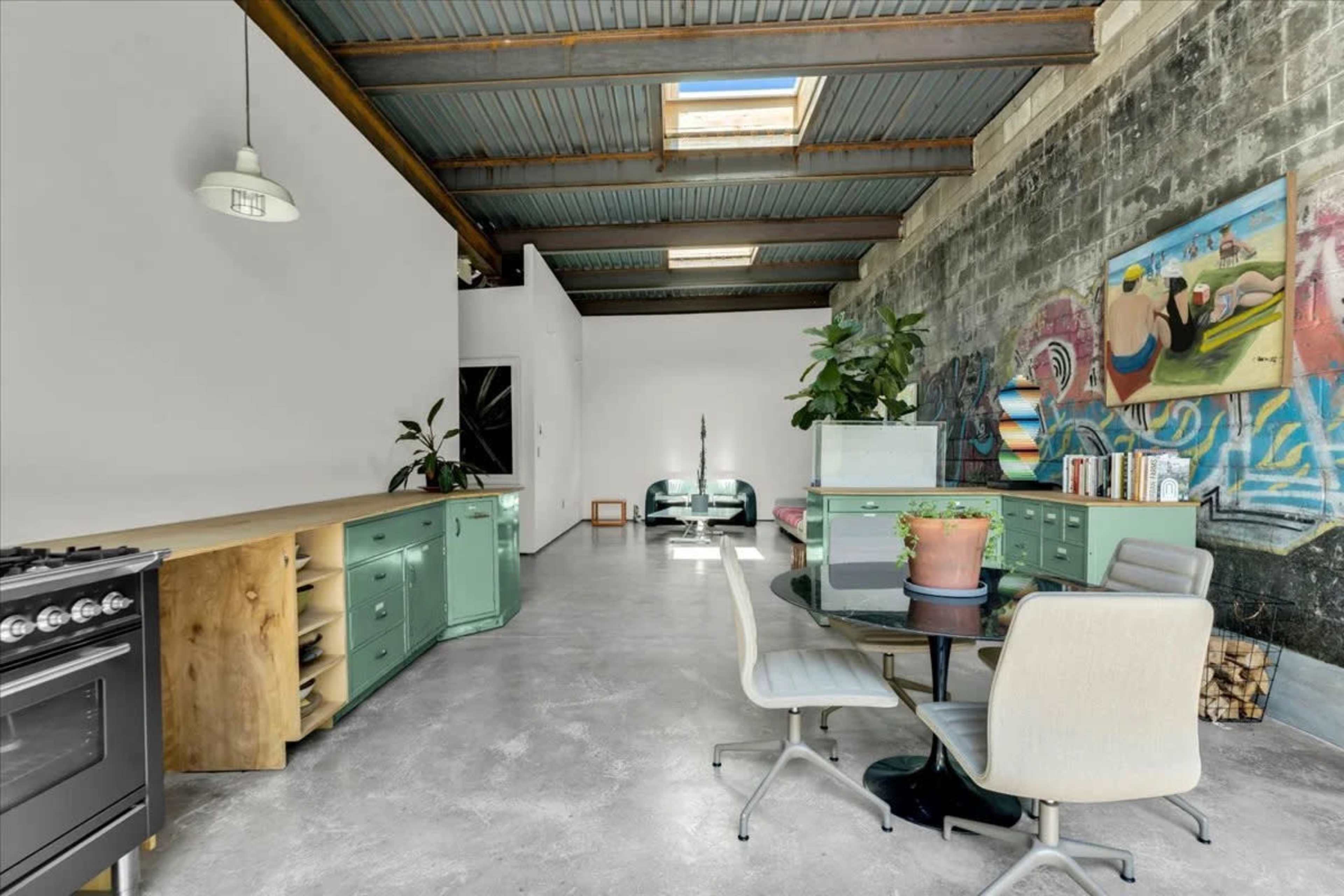 The image shows an industrial-style living space featuring a kitchen area with green cabinets, a dining table, and a seating area adorned with plants and artwork on the walls.