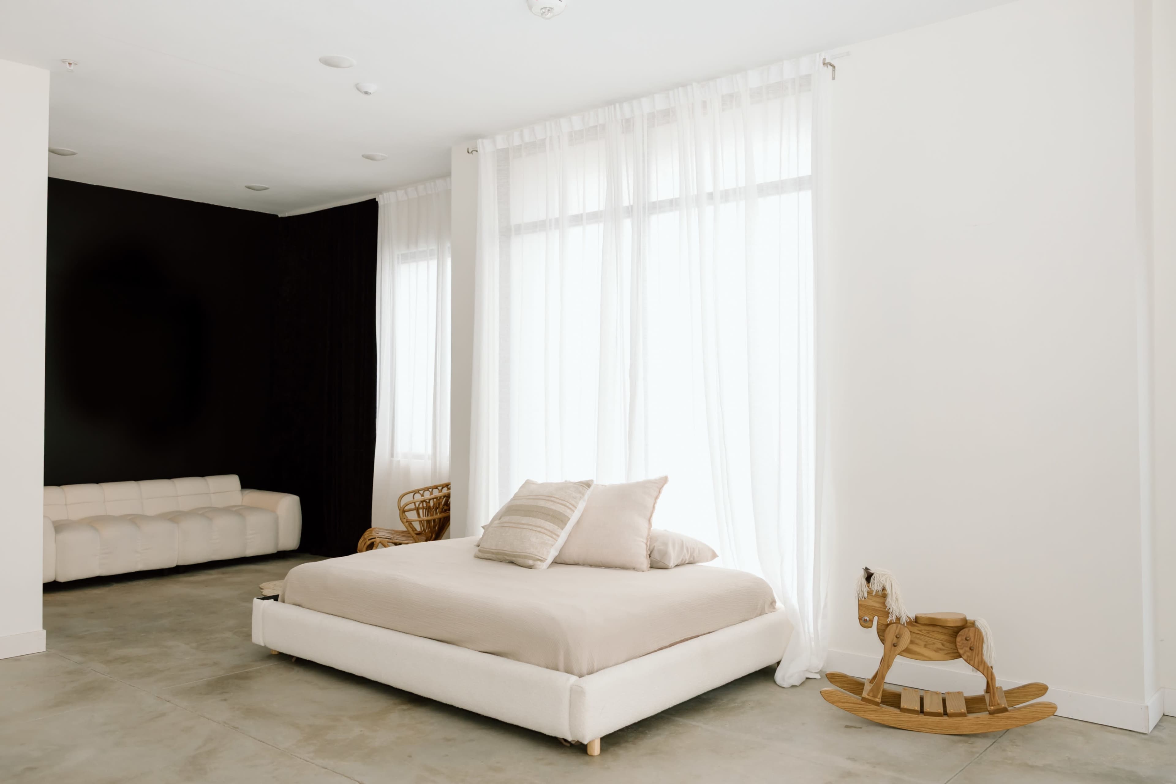 The image shows a minimalistic bedroom featuring a low-profile bed with light bedding, large windows covered by sheer white curtains, and a wooden rocking horse in the corner.