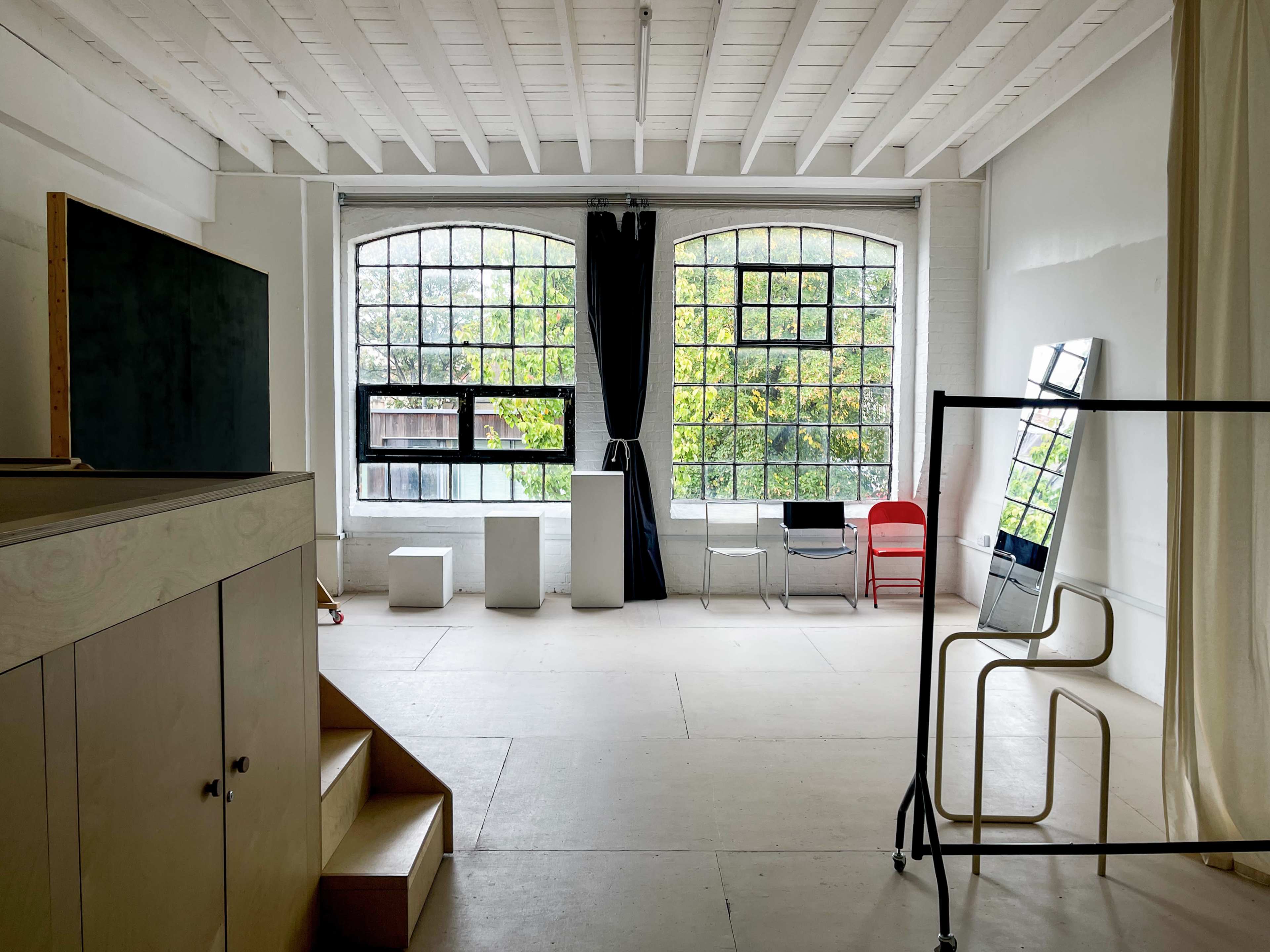 The image depicts a spacious, minimalistic interior with large windows, a staircase, and various furniture pieces, including a mirror and chairs.