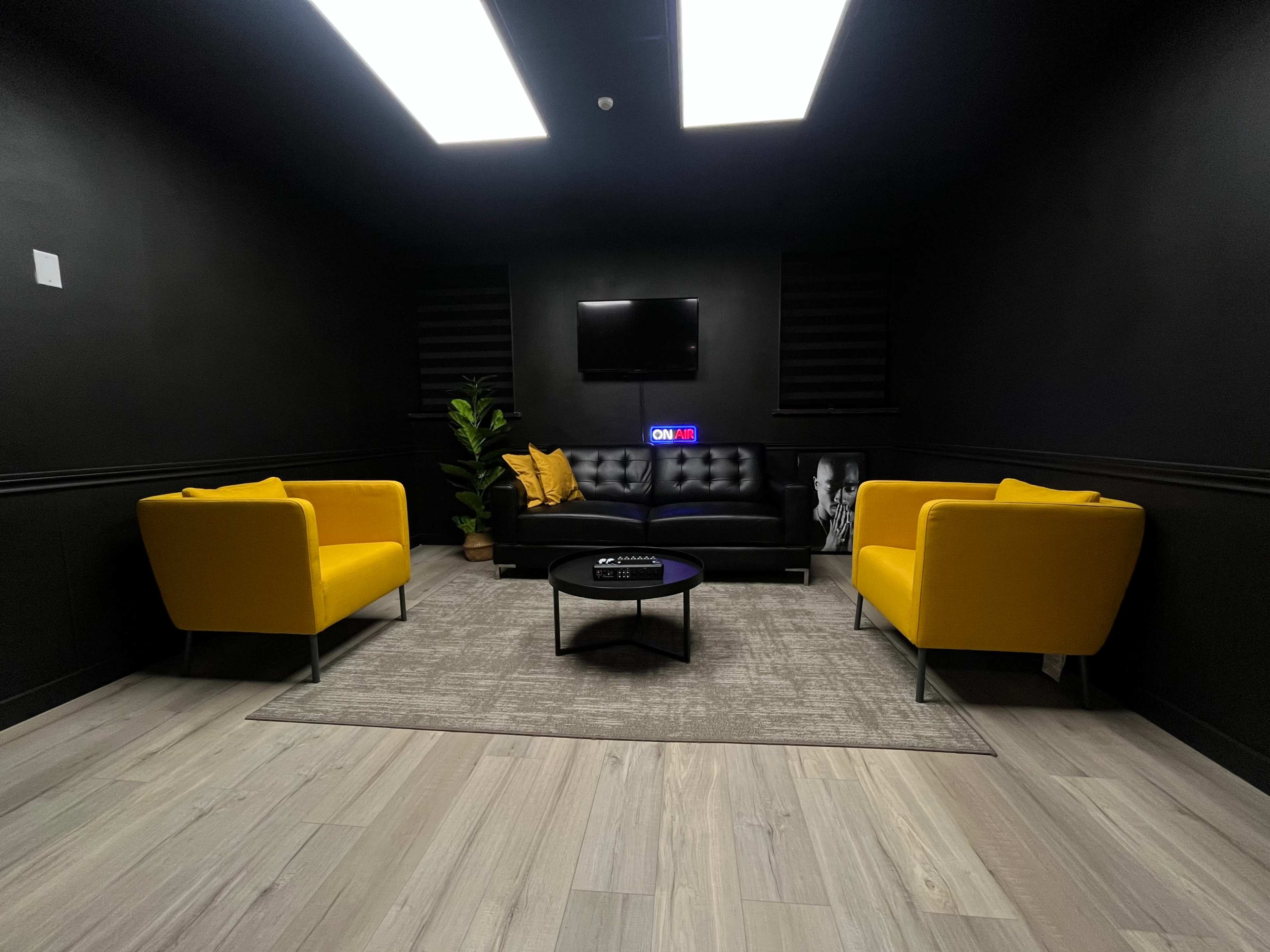 A black-walled room featuring two yellow chairs and a black sofa, with a coffee table in the center and a television mounted on the wall.
