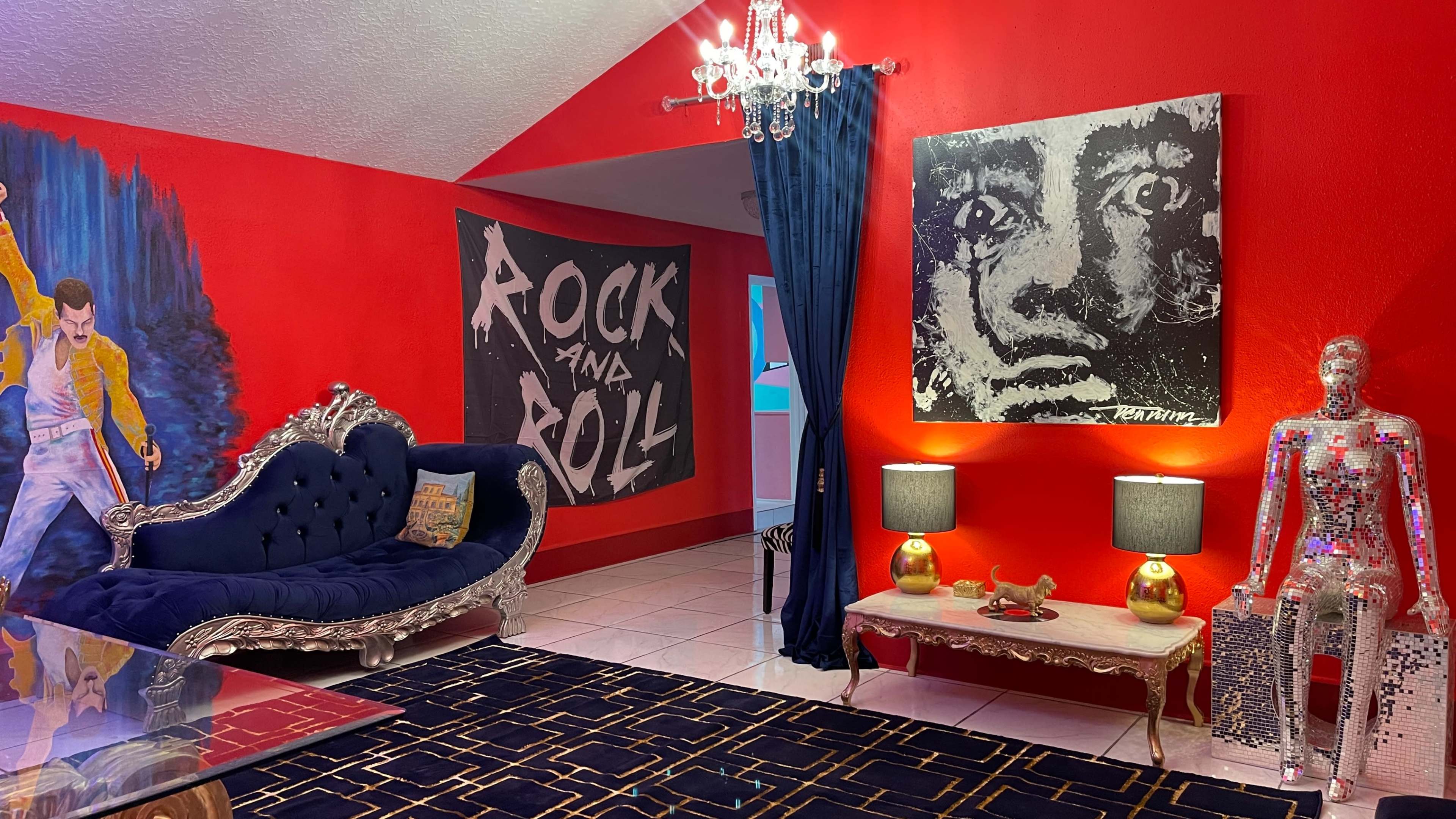 The room features a red wall with artwork, a blue velvet sofa, a table with a mirrored base, and two lamps flanking a metallic figure.