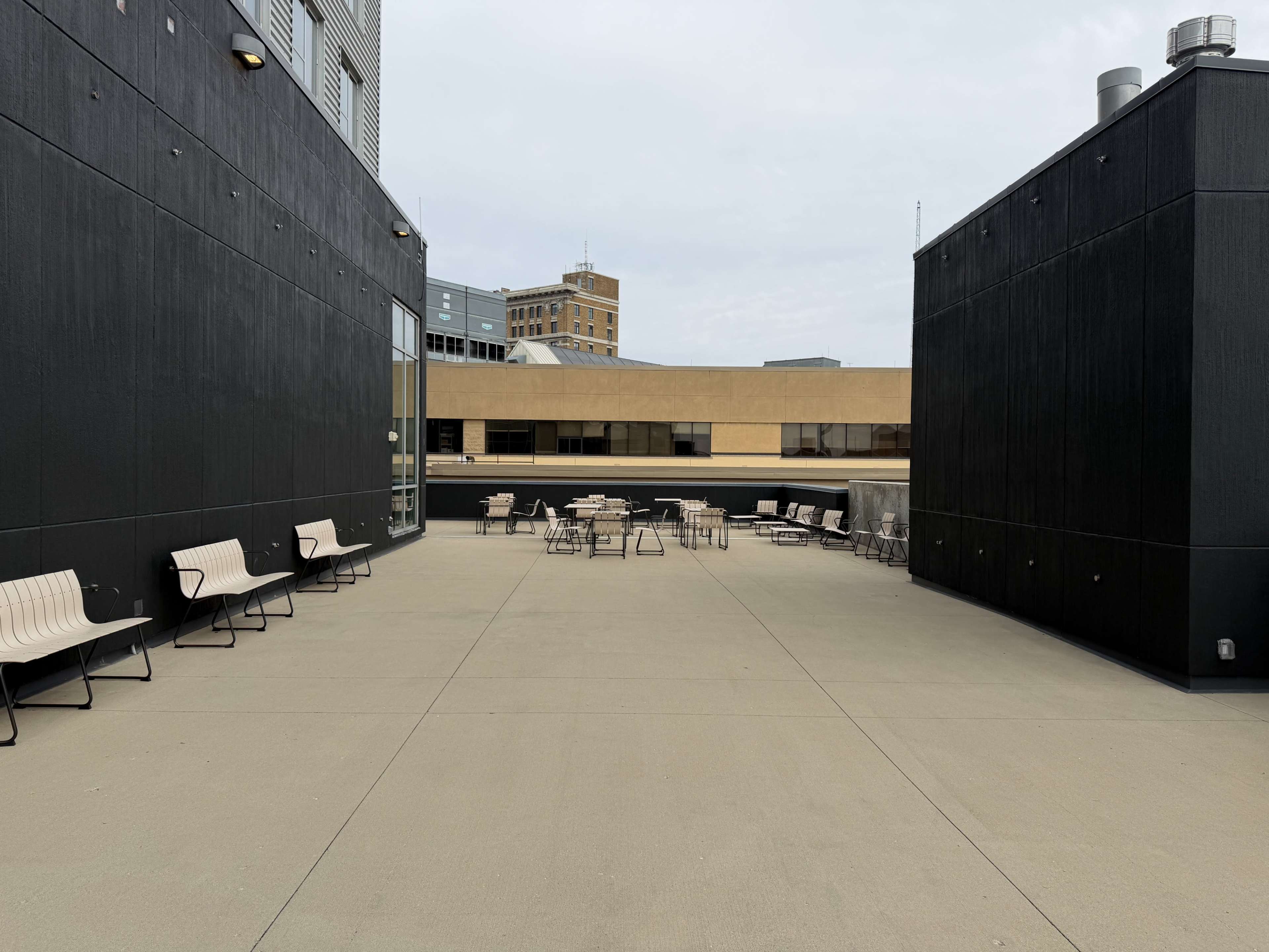The image shows a rooftop terrace with several tables and chairs surrounded by tall, dark walls and buildings in the background.