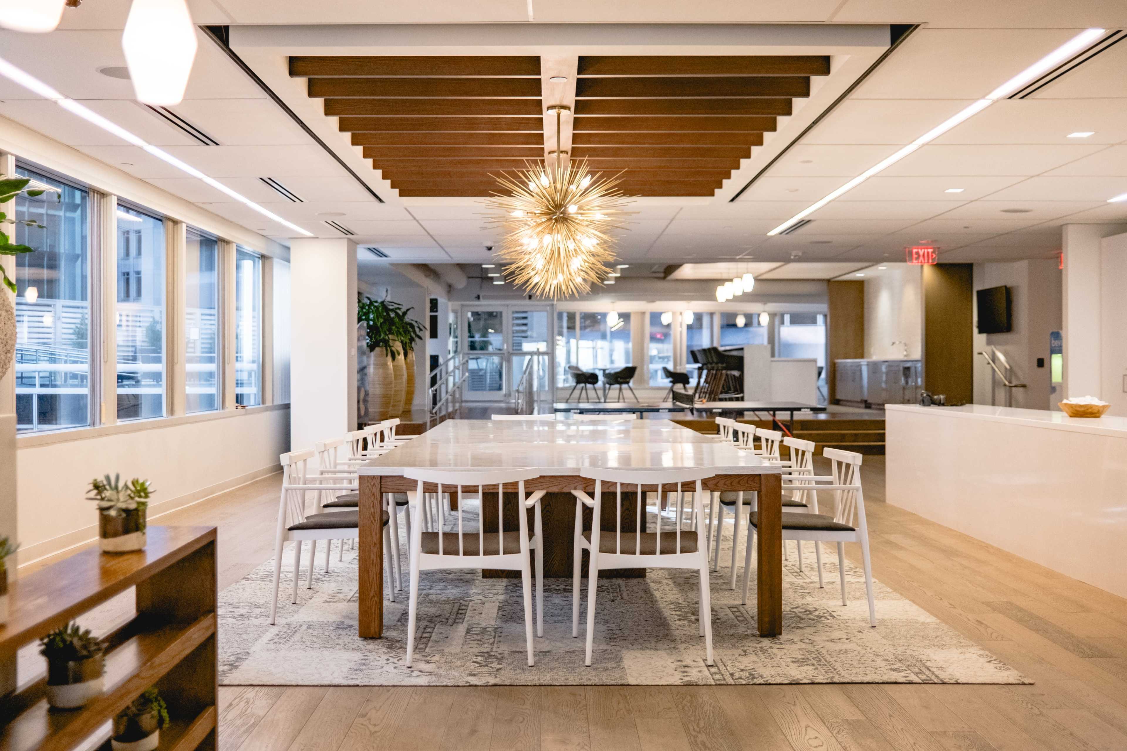 A modern dining area features a large wooden table surrounded by white chairs, with a striking light fixture hanging above and large windows providing ample natural light.