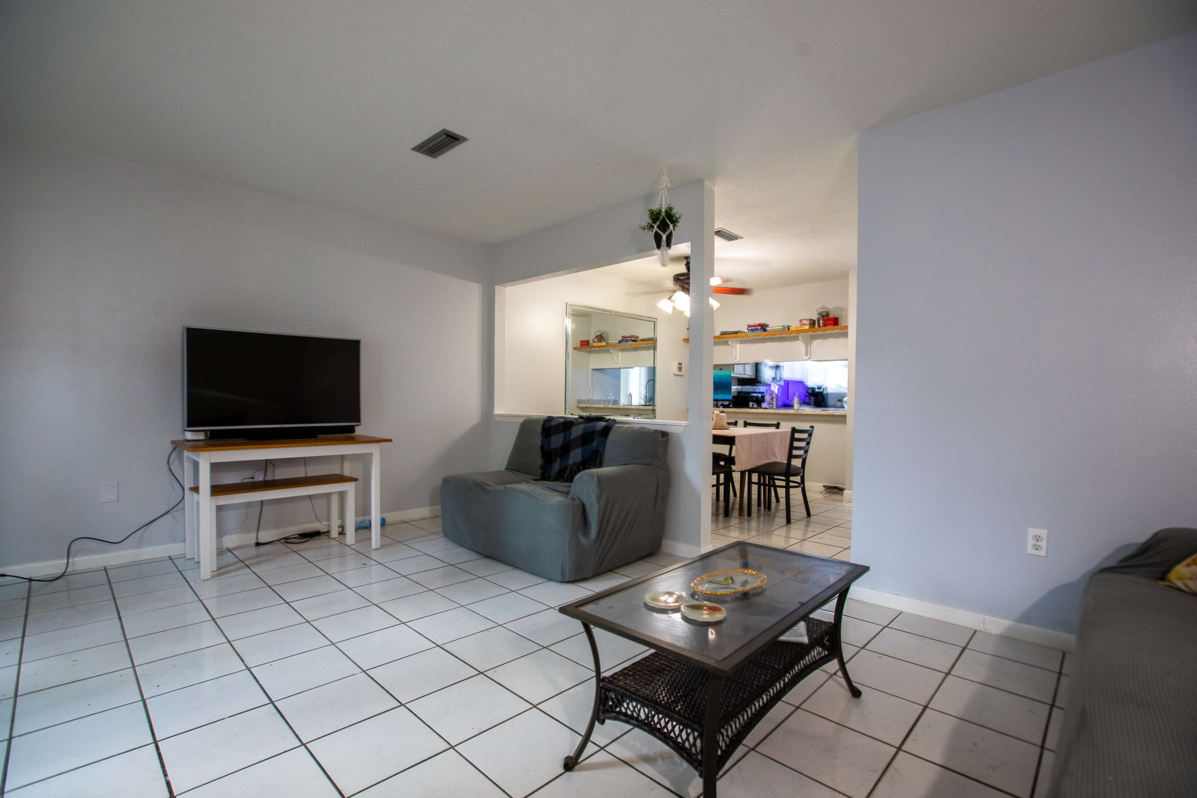 A living room features a television on a white stand, a gray sofa, and a glass-top coffee table, with a view of a kitchen in the background.