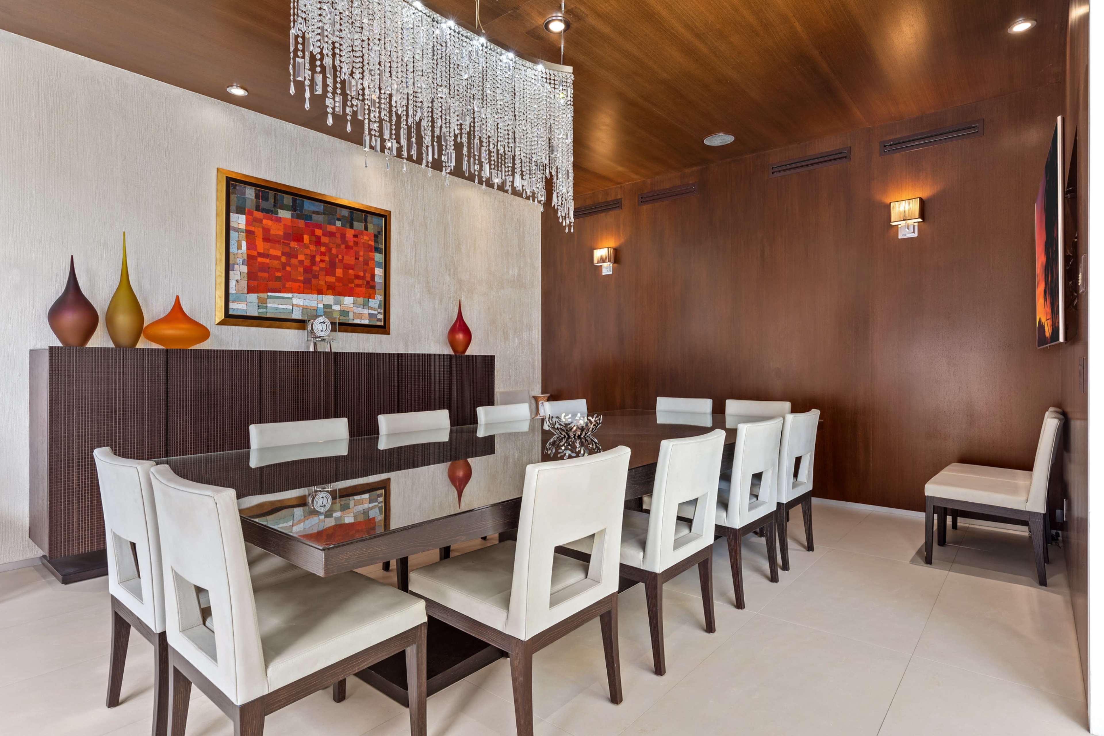 A modern dining room features a large dark wooden table surrounded by white chairs, with a decorative chandelier above and minimalist artwork on the walls.