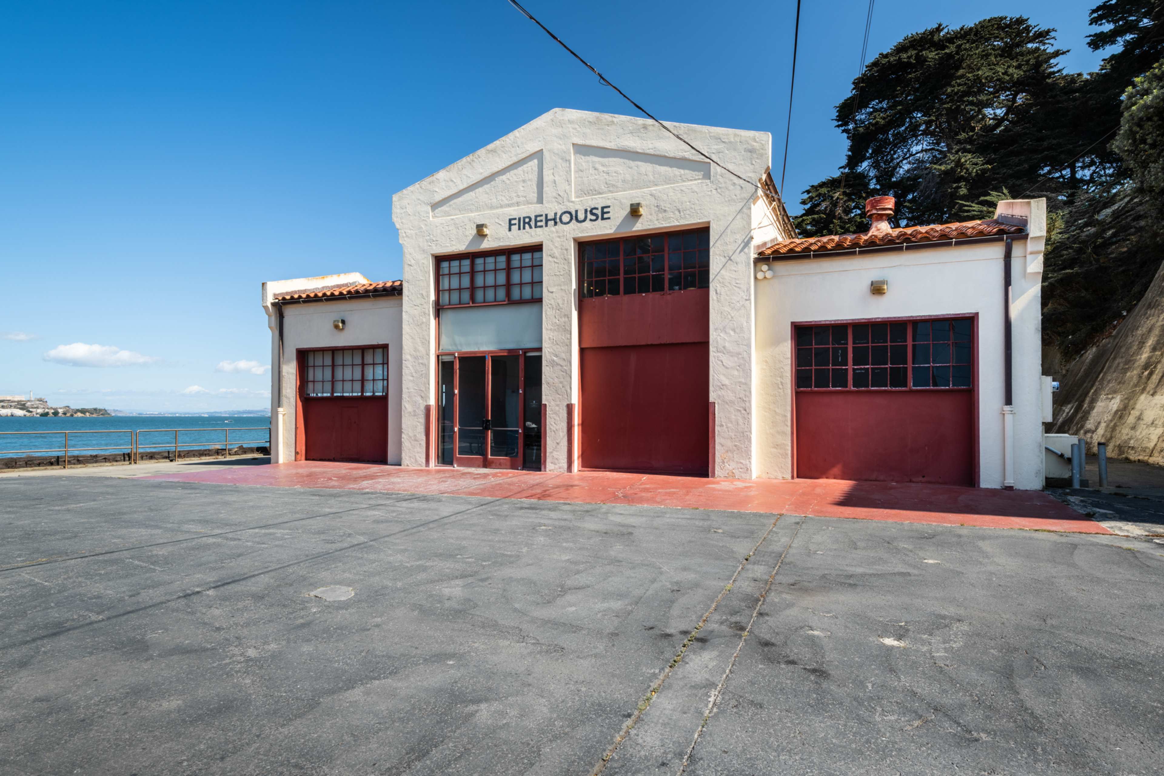 An old firehouse building with large red doors, located beside a calm waterfront under a clear sky.