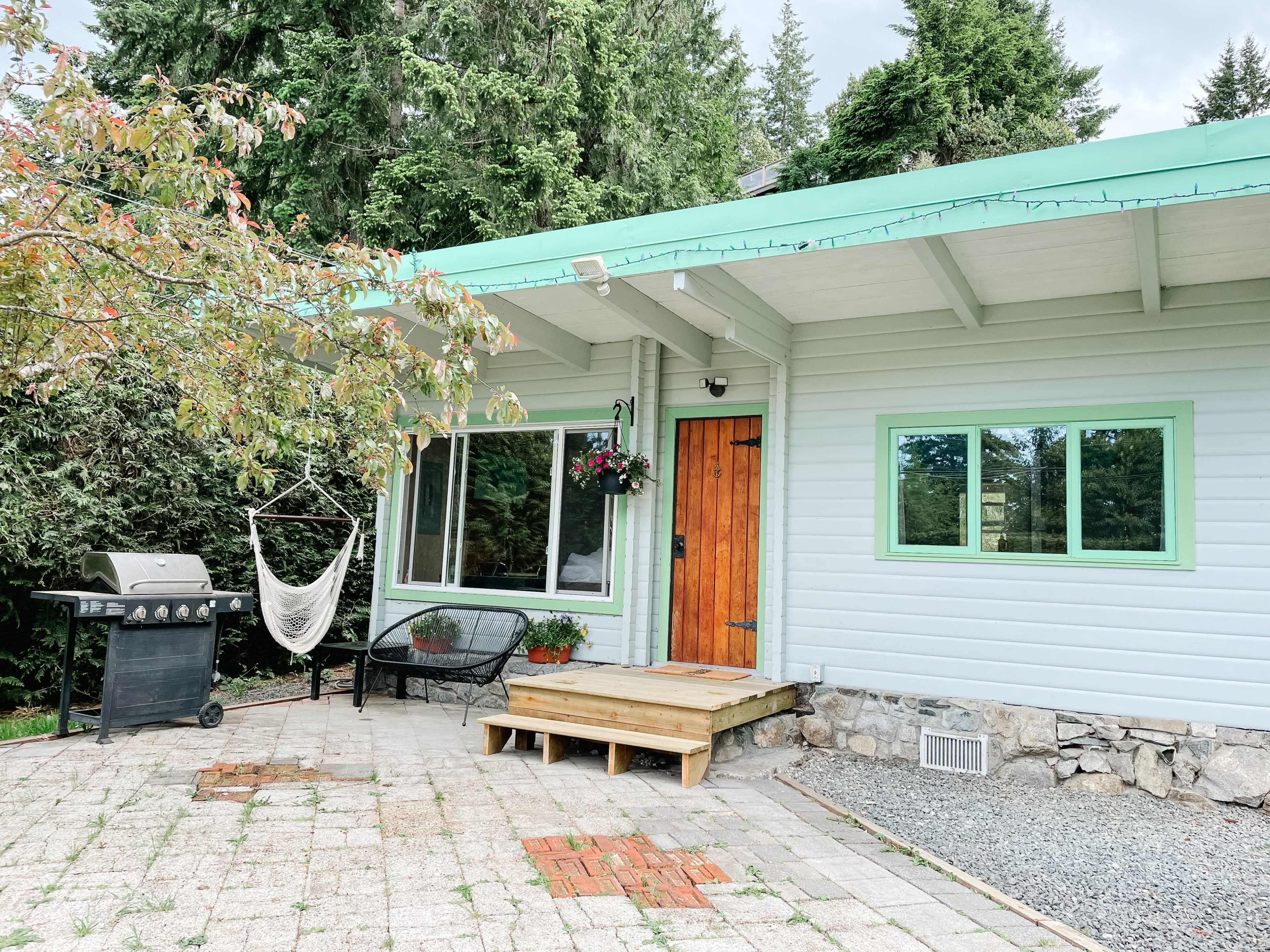 A rustic house with a wooden door, a patio area featuring a grill and a hammock, surrounded by green trees.