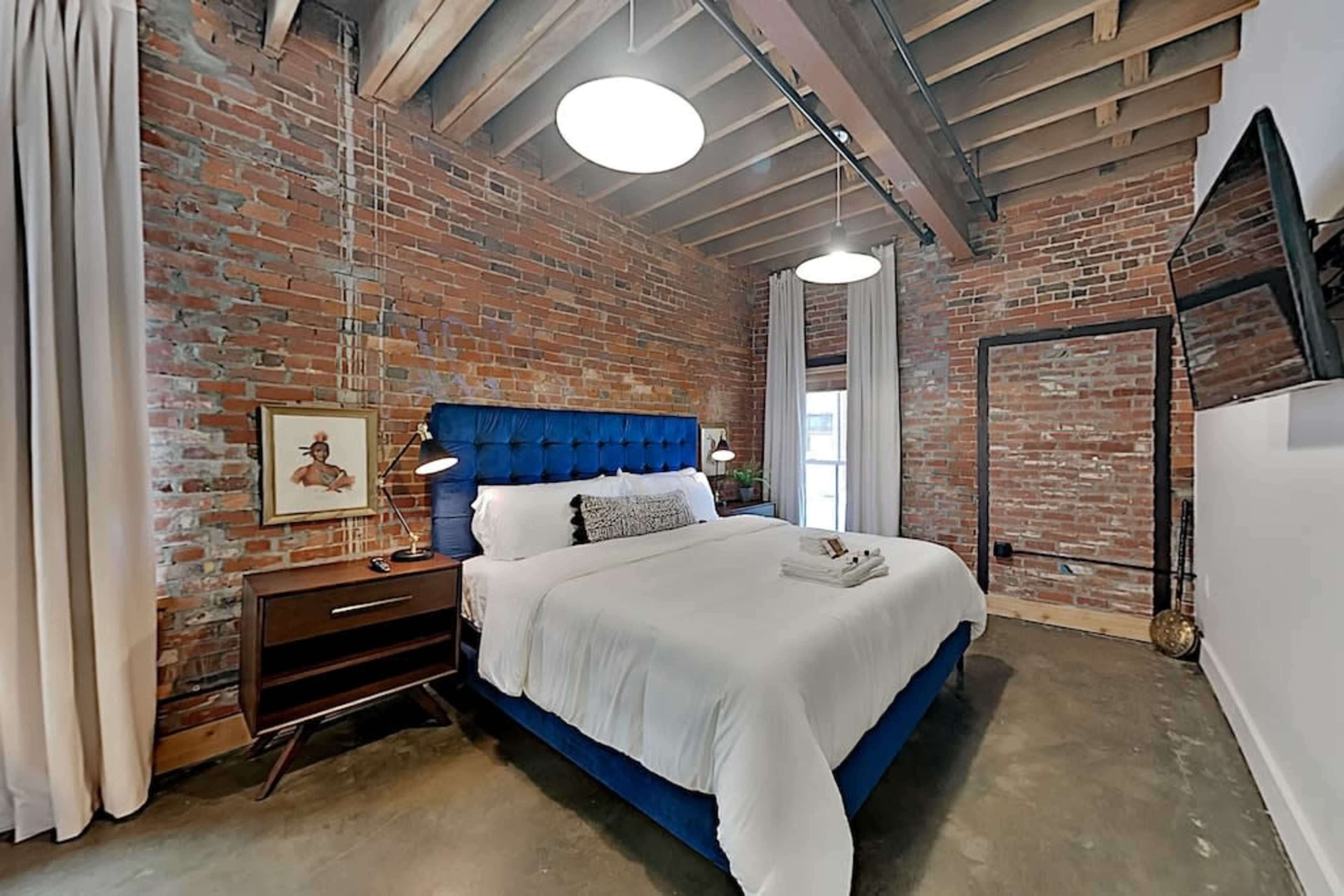 The bedroom features exposed brick walls, a blue upholstered bed, and large windows with sheer curtains.