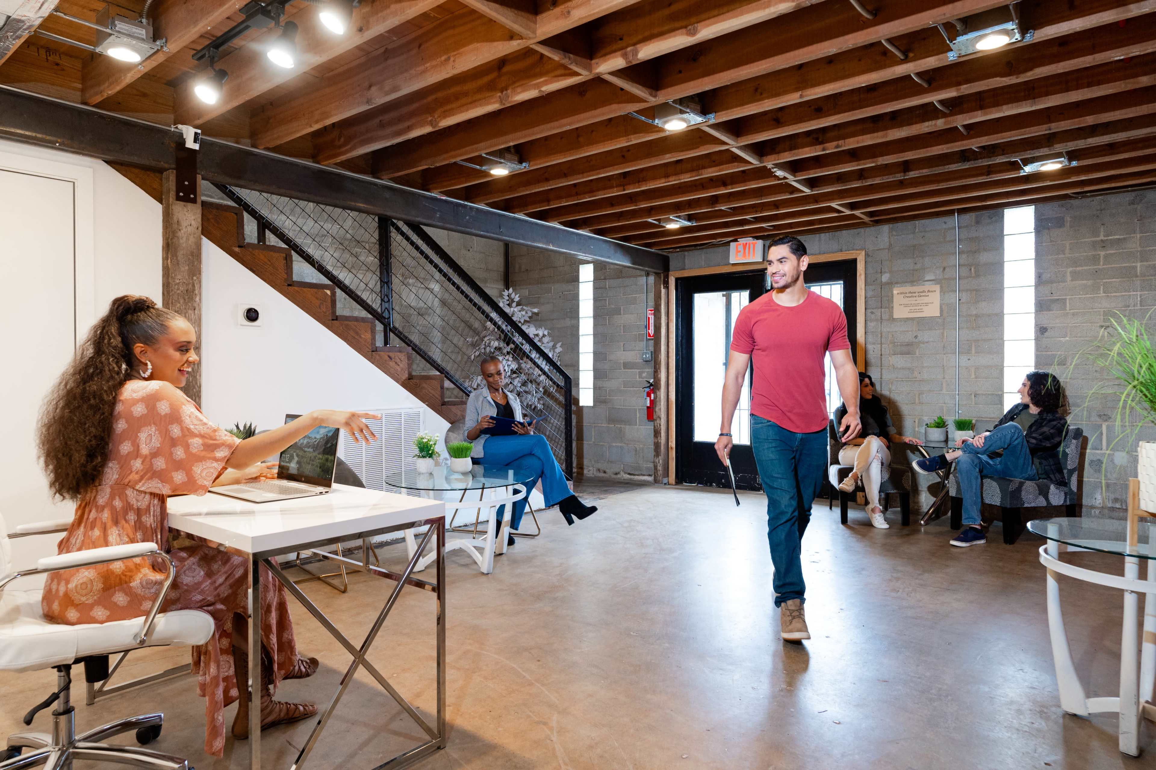 A man walks through a modern office space with several people seated and a staircase in the background.