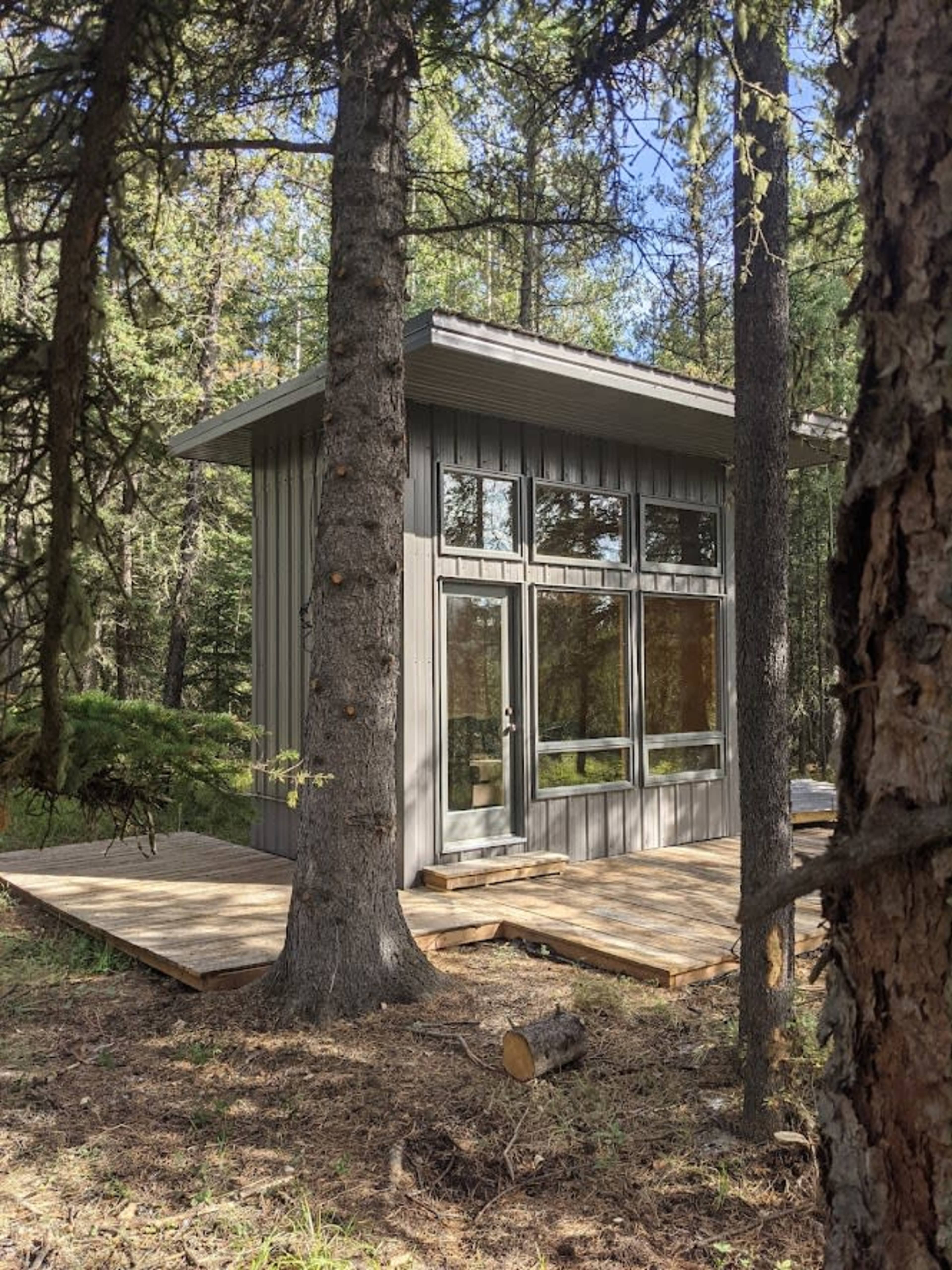A modern cabin with large windows is set on a wooden deck among tall trees in a forested area.