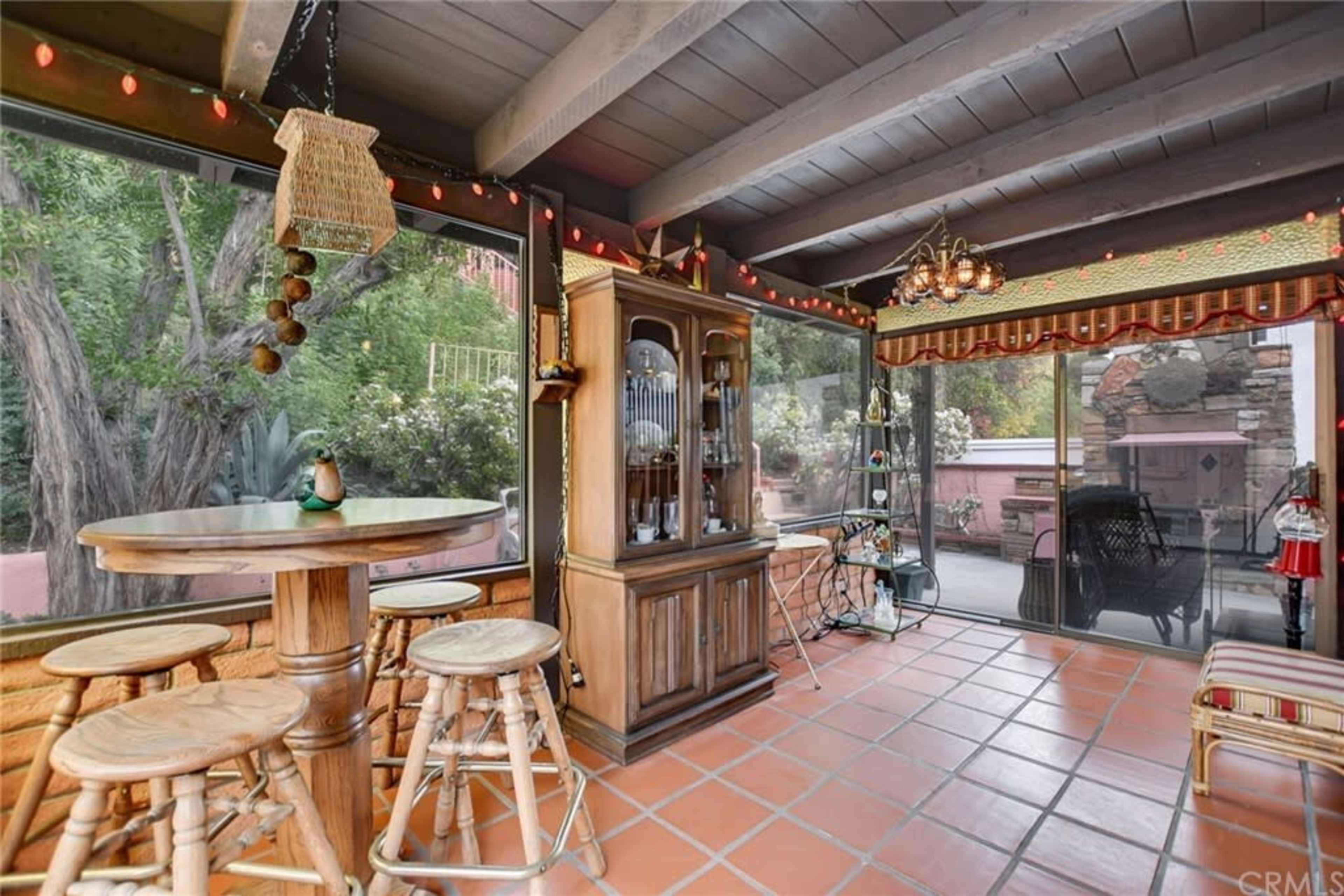 The image shows a screened-in patio featuring wooden furniture, a cabinet with glass display, and decorative lights along the ceiling.