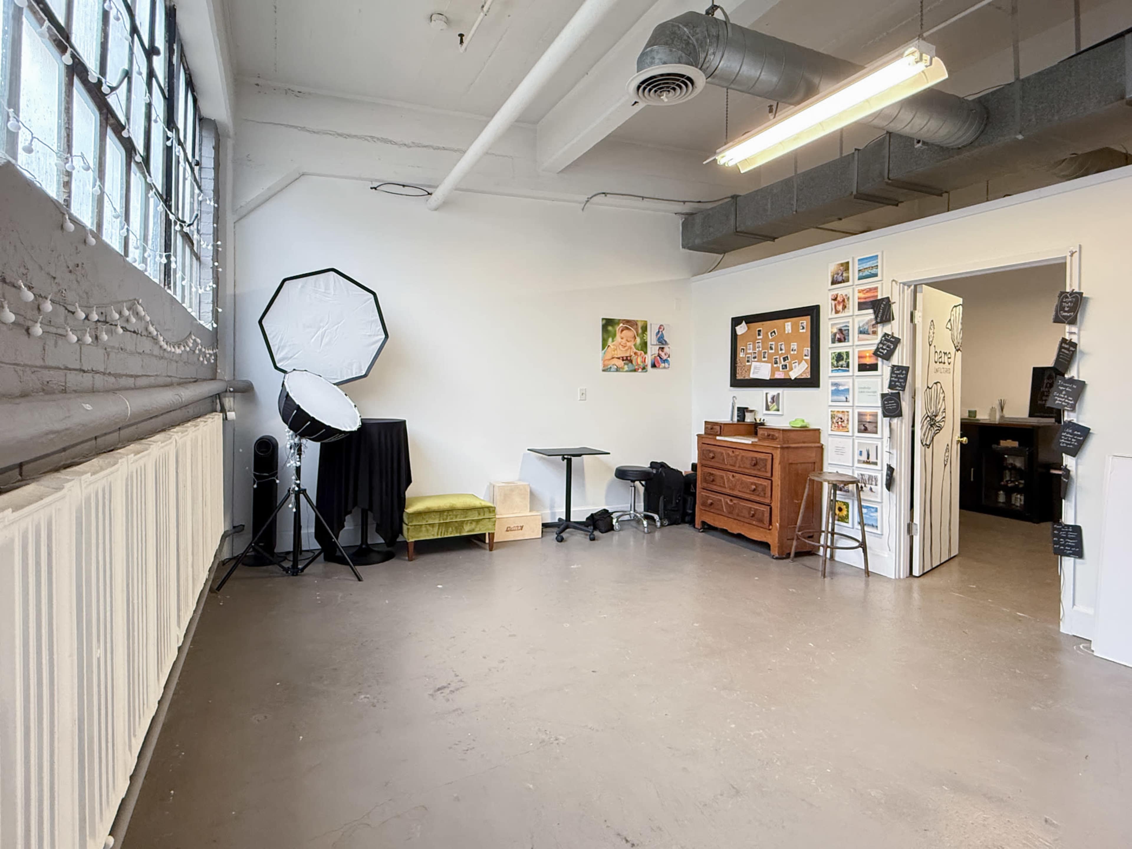 The image shows a spacious, empty studio with large windows, a green chair, a wooden dresser, and a backdrop stand with lighting equipment.