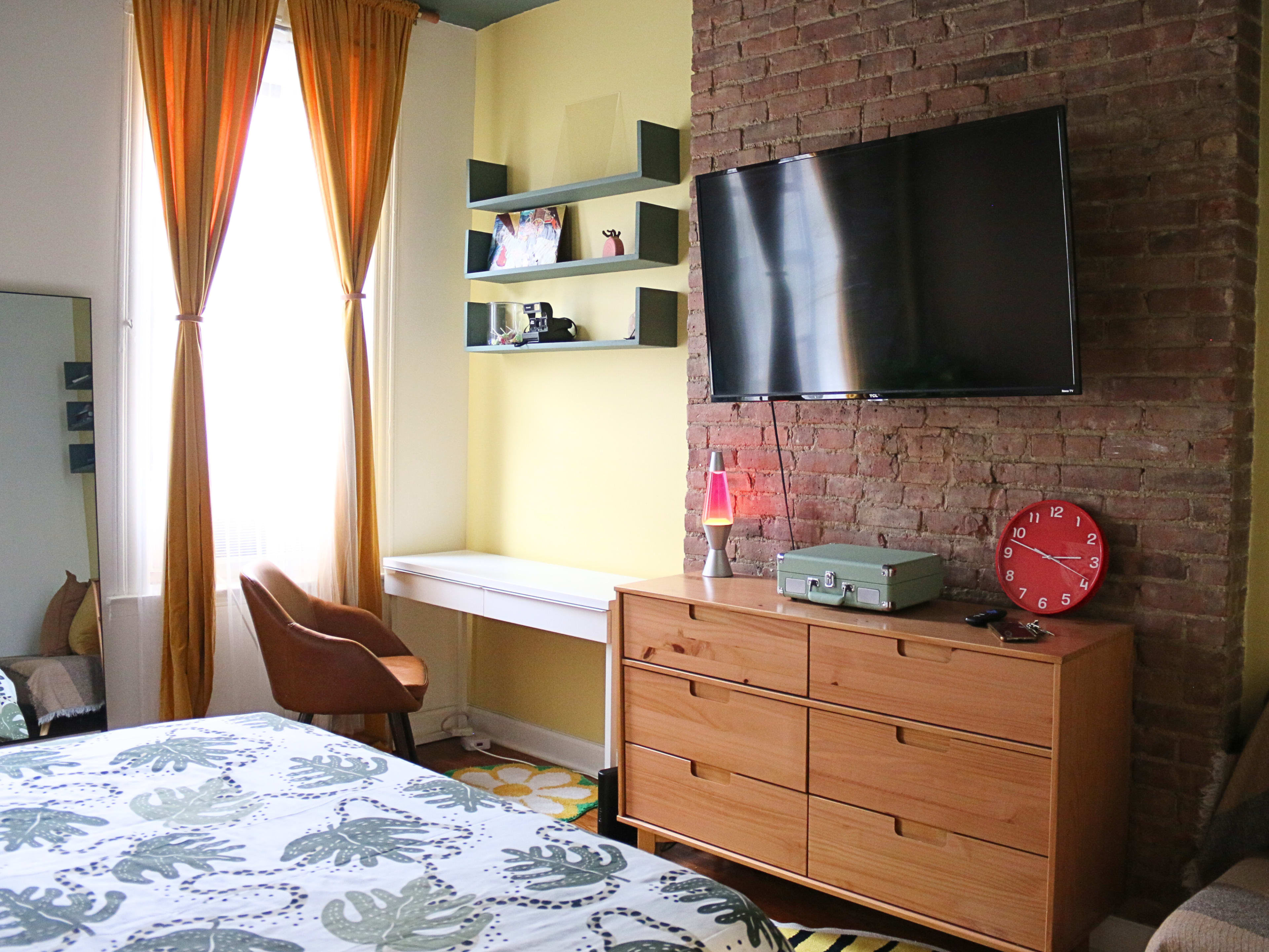 A cozy bedroom featuring a brick wall, a television mounted on the wall, a wooden dresser, a desk with shelving above, and a chair beside a large window adorned with yellow curtains.