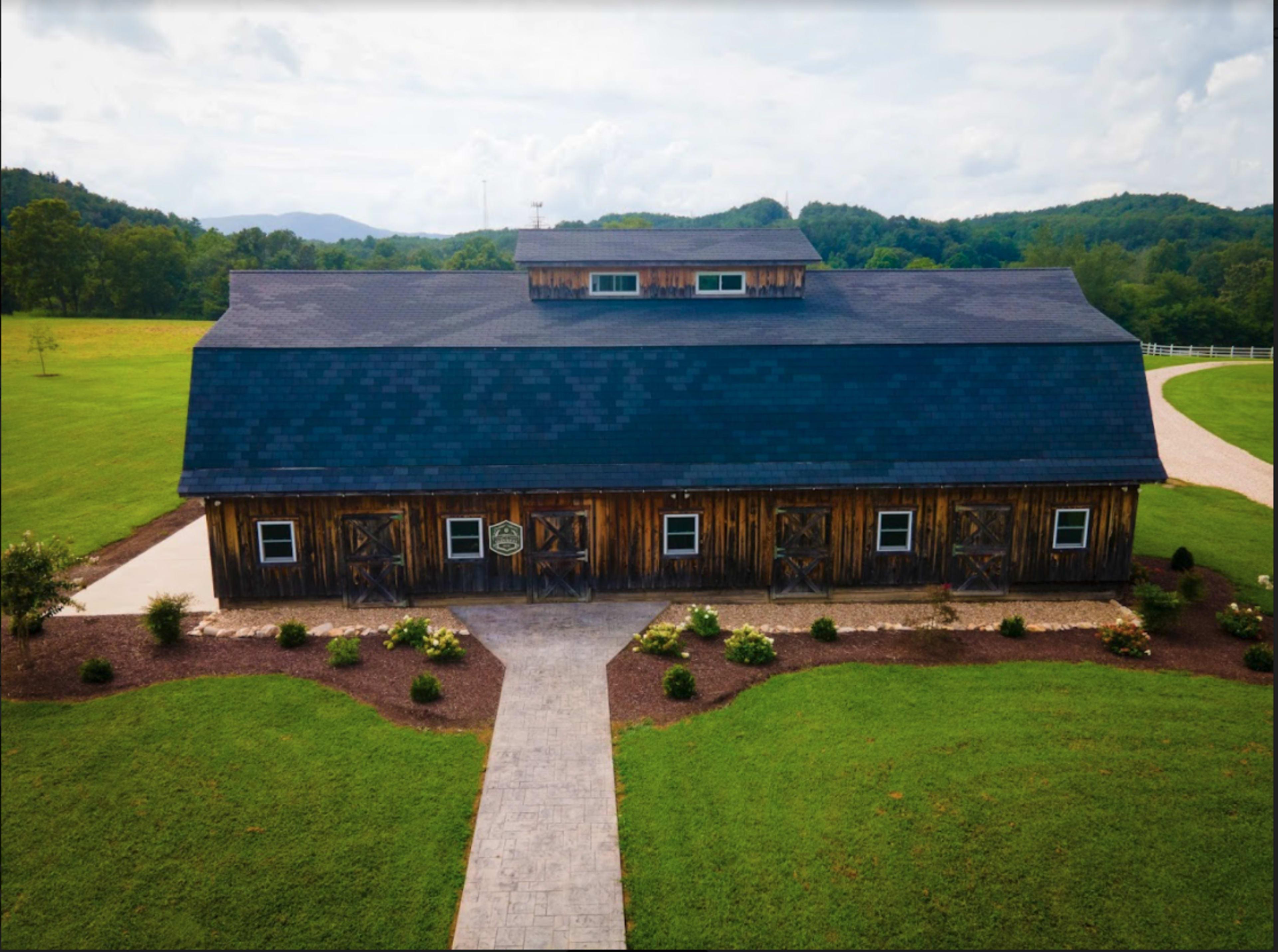 A large, wooden barn with a dark roof situated on a green lawn, surrounded by trees and shrubs.