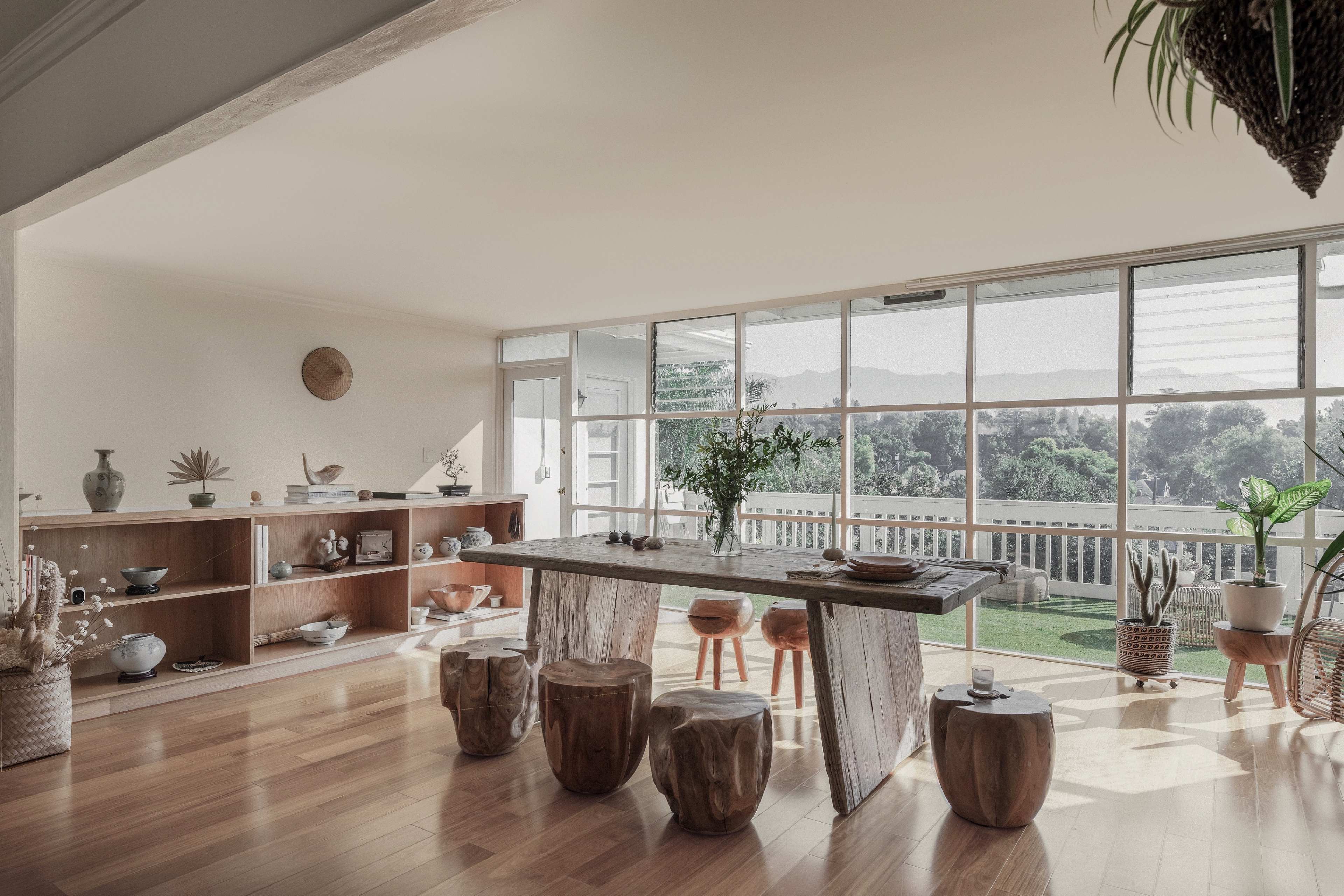 A spacious, well-lit room features a wooden dining table surrounded by tree stump stools, with shelves displaying various ceramic items and a view of greenery through large glass windows.