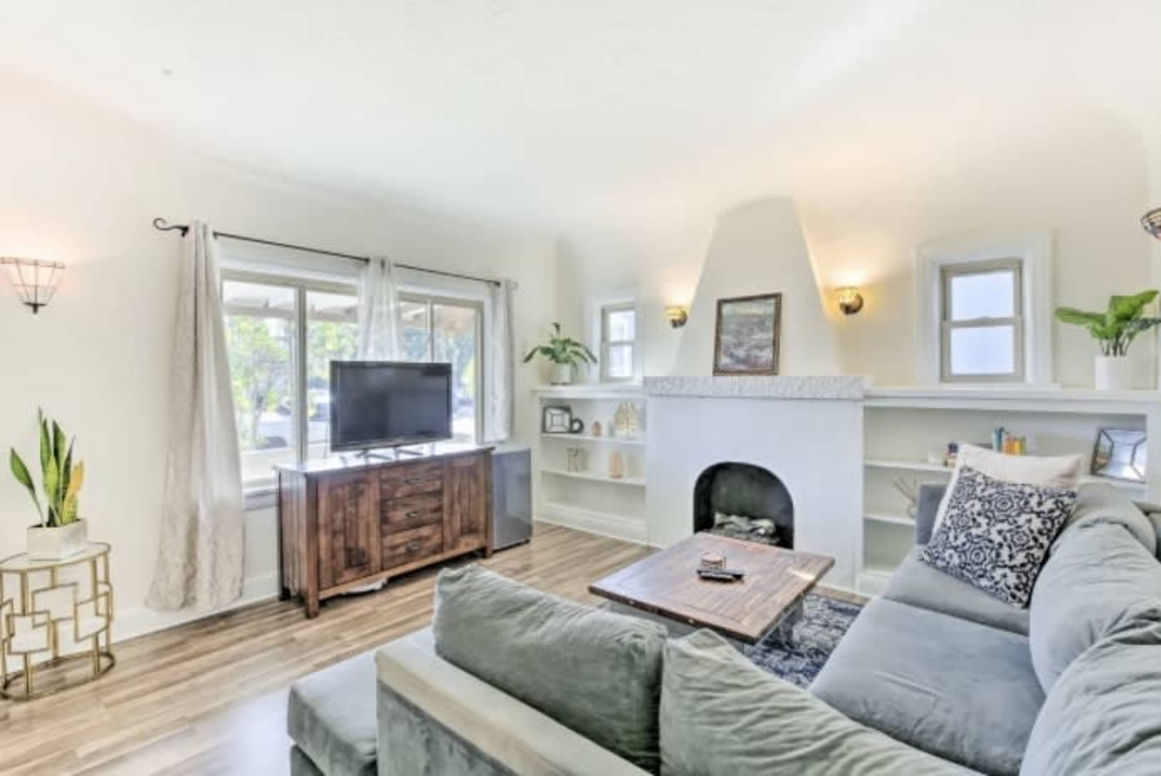 The living room features a sectional sofa, a wooden TV stand, and a large window with natural light illuminating the space.