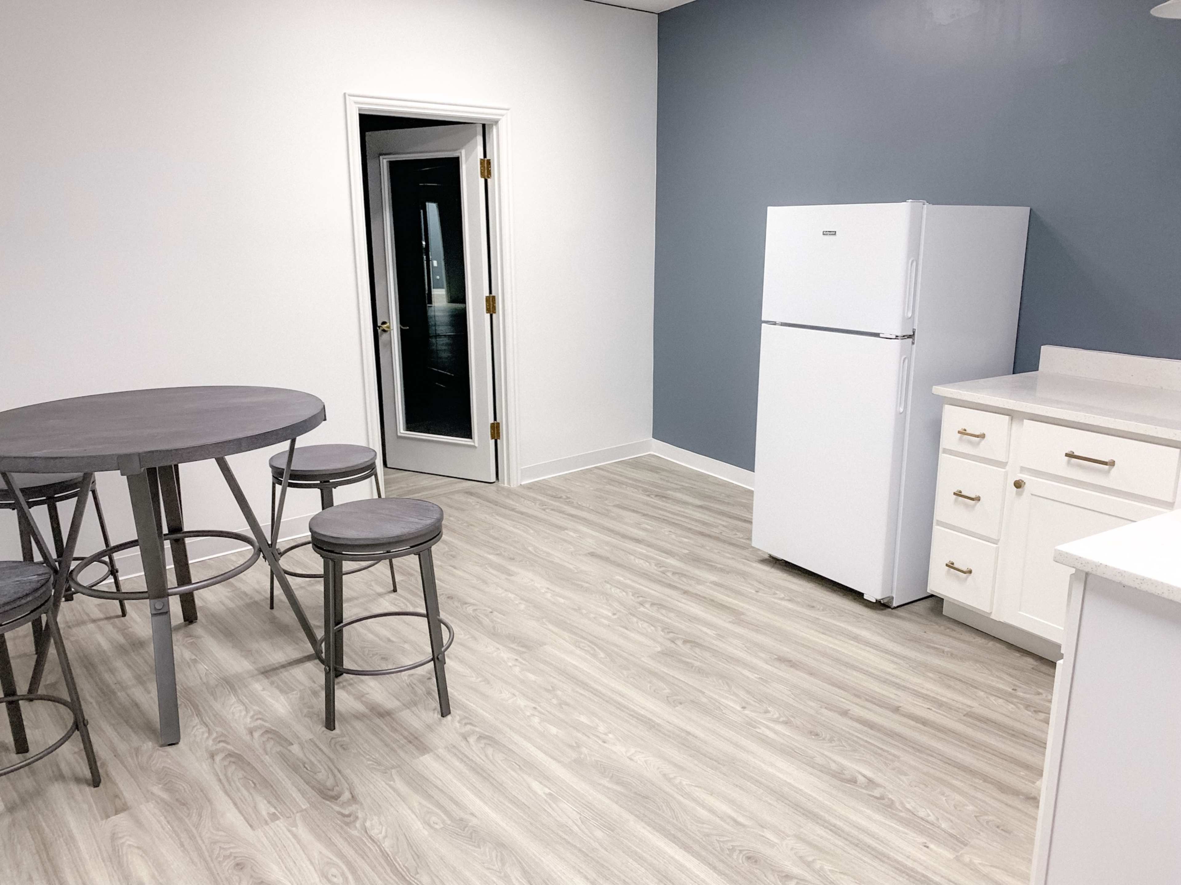 A simple kitchen area featuring a white refrigerator, a round dining table with stools, and a gray wall.