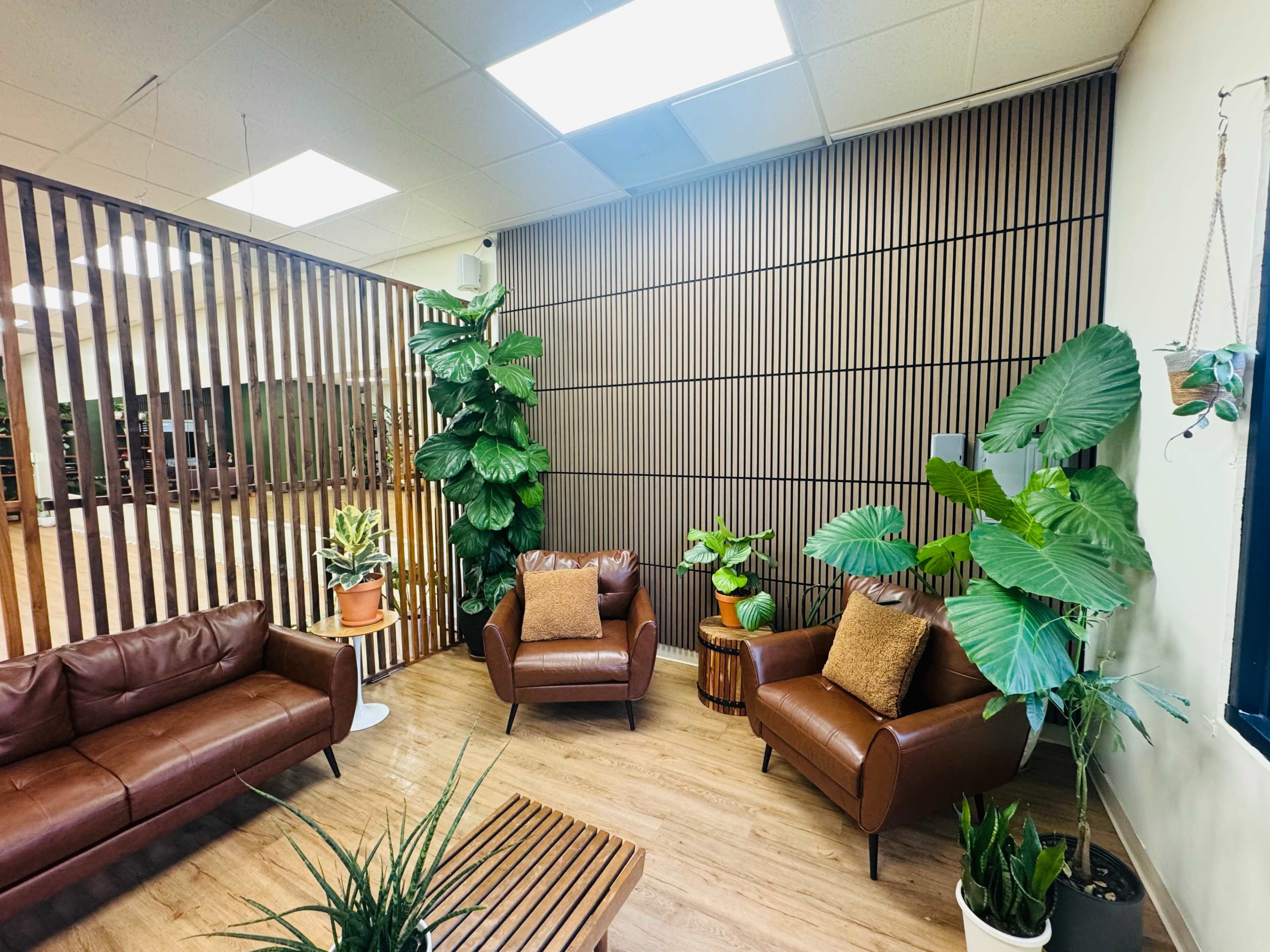 The image shows a cozy waiting area furnished with brown leather sofas, wooden accents, potted plants, and wooden flooring beneath bright overhead lights.