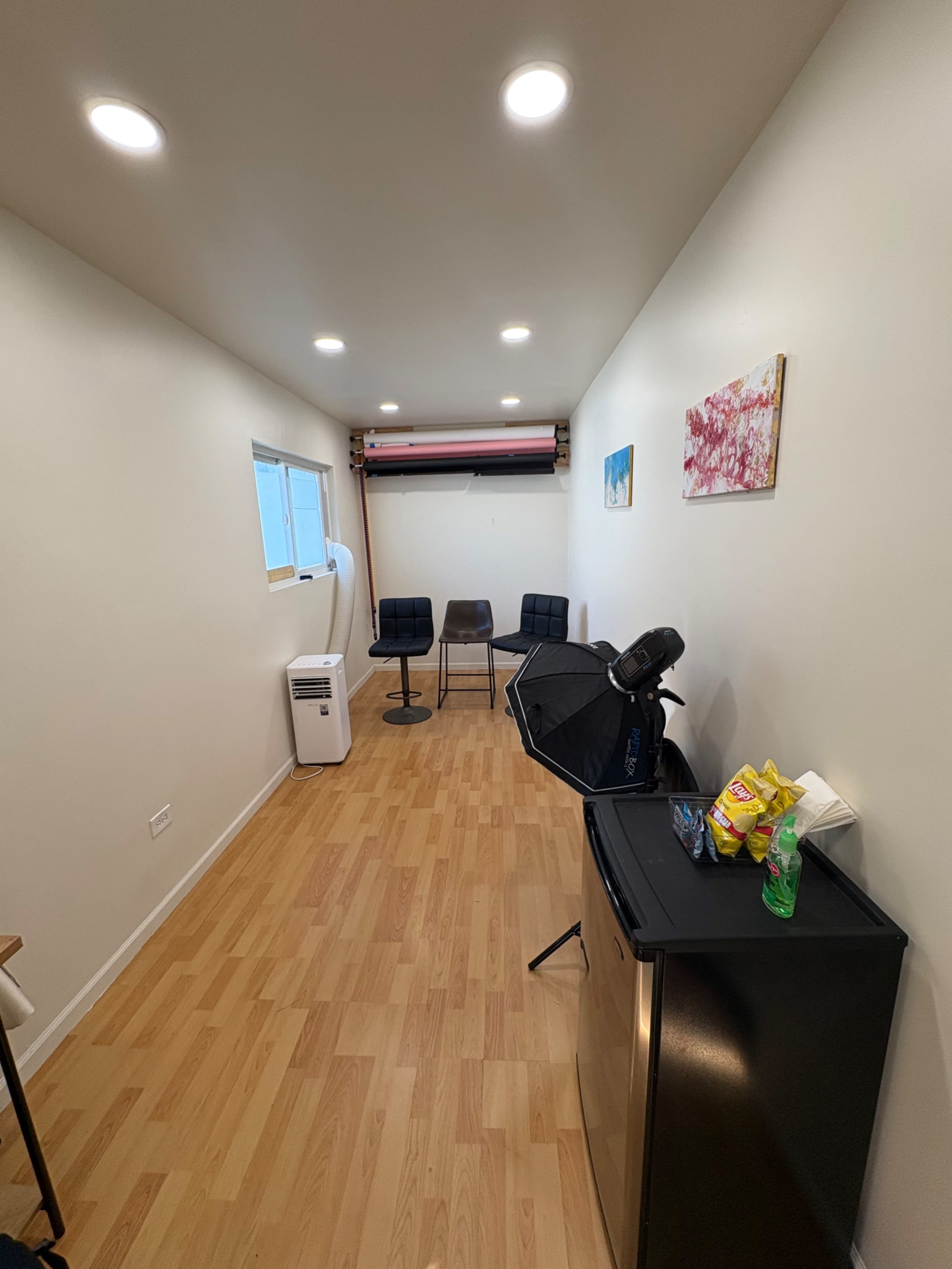 The image depicts a narrow room with wooden flooring, featuring a small refrigerator, chairs, a light setup, and a window along one wall.