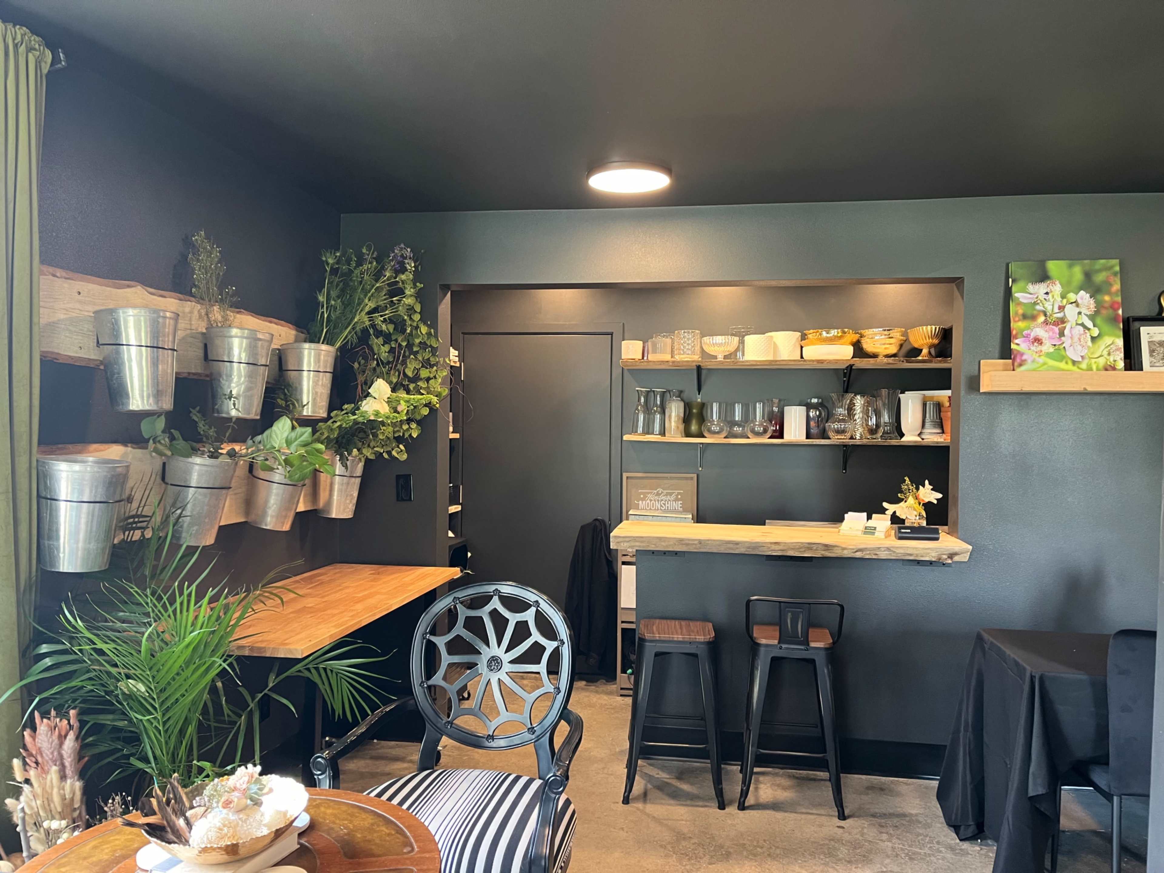 A cozy café interior with a wooden table, metal chairs, and shelves displaying potted plants and dishware against dark walls.