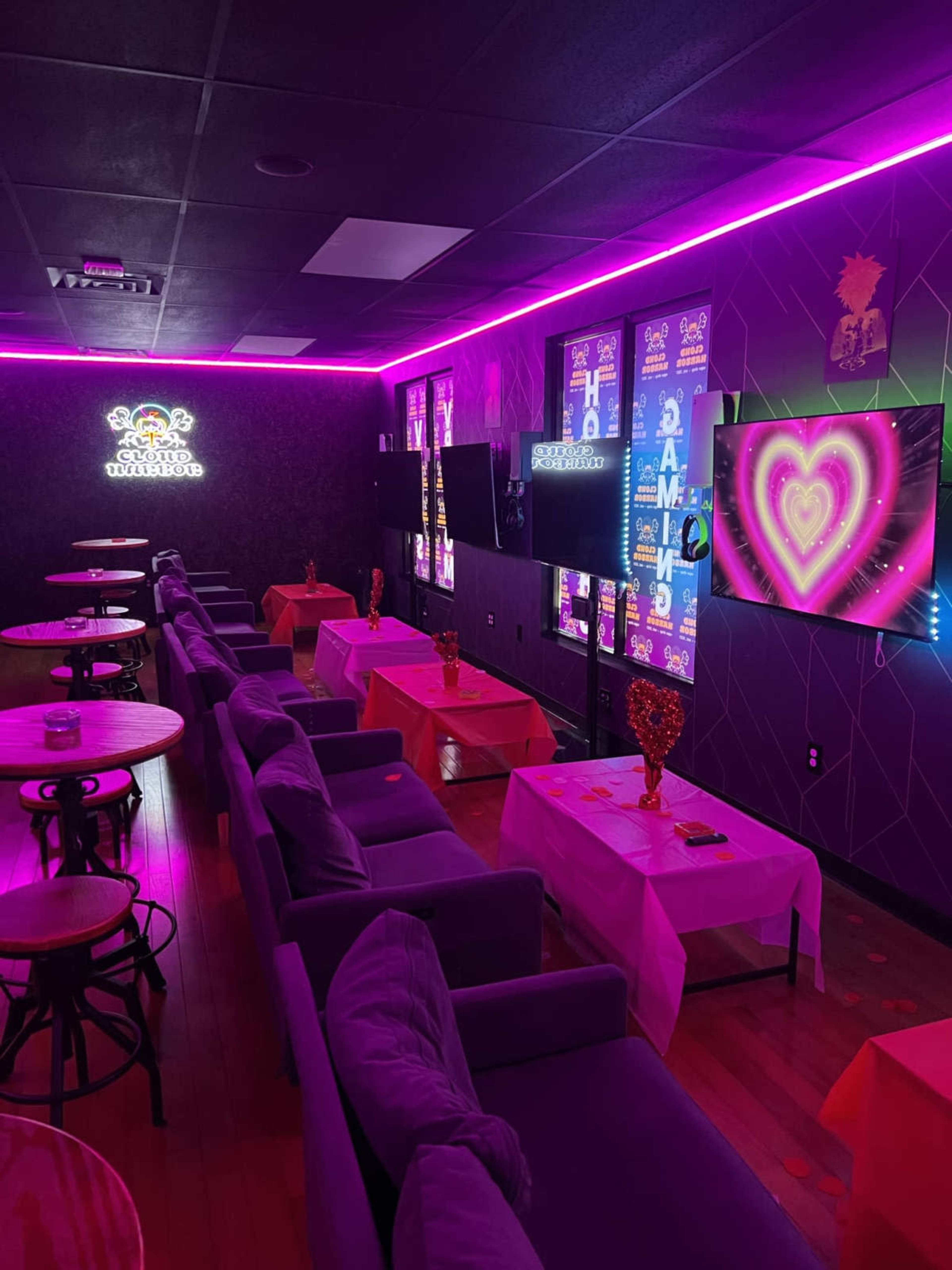 The image shows a dimly lit lounge area with purple neon lights, featuring several tables and chairs arranged in front of wall-mounted televisions.