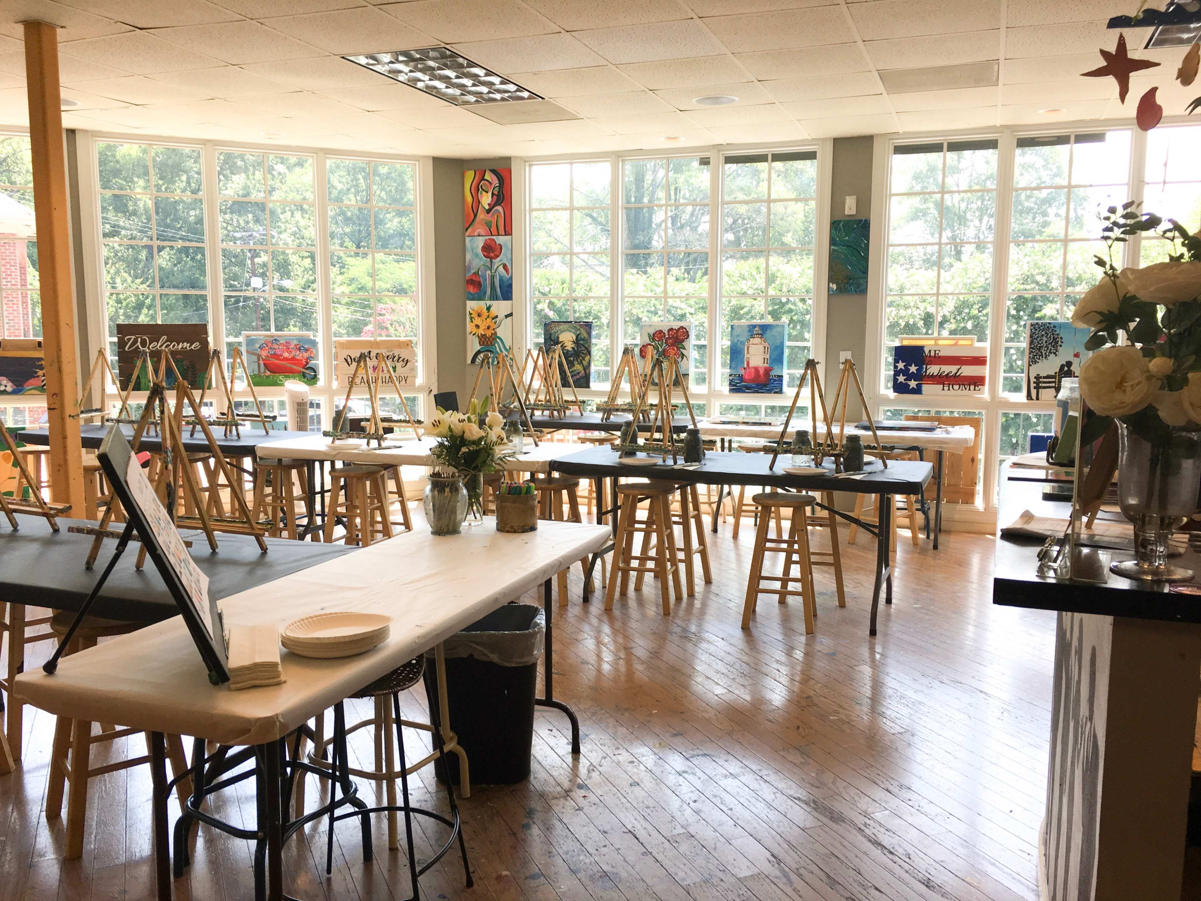 An art studio with numerous easels set up around tables, large windows letting in natural light, and artwork displayed on the walls.