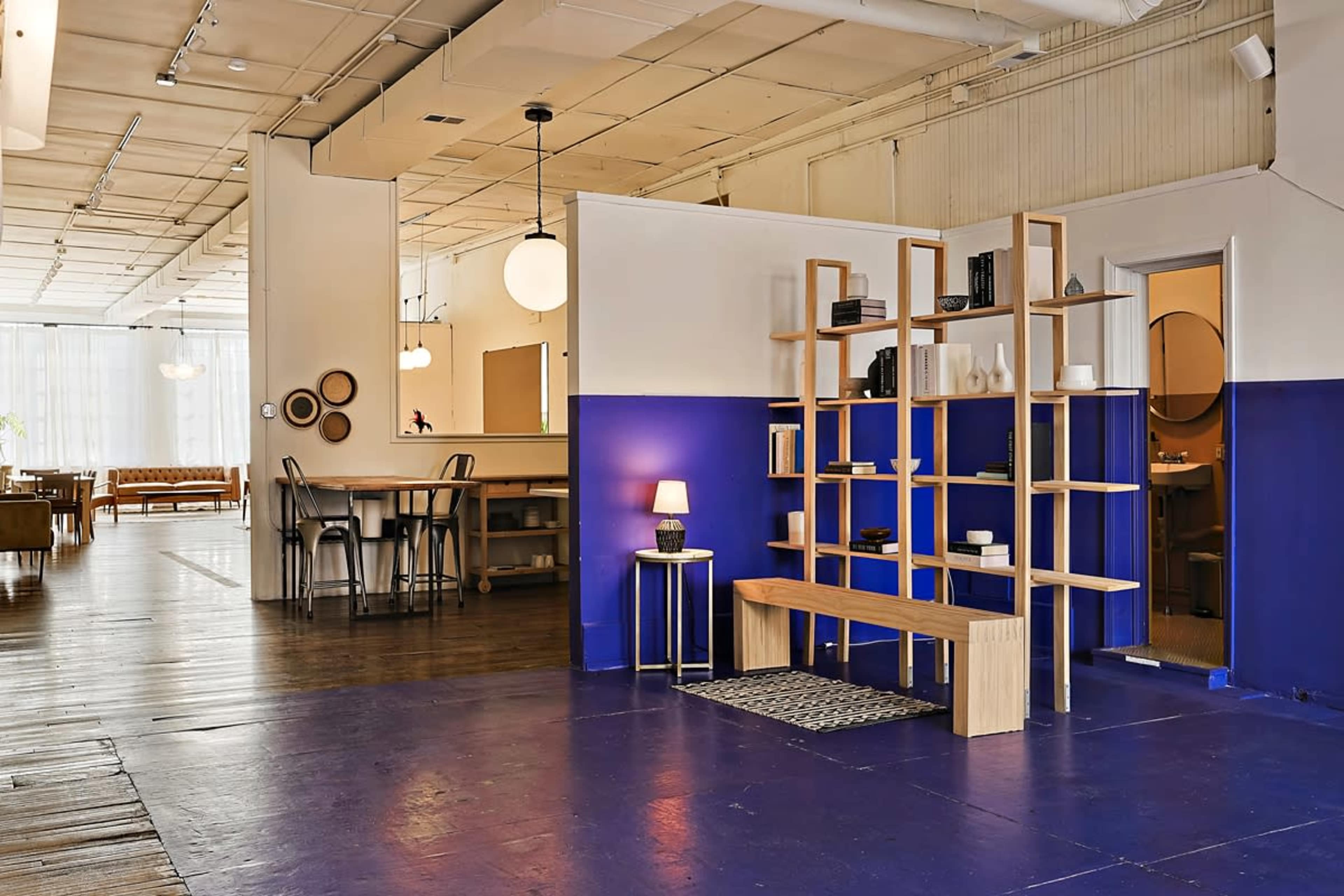 The image shows a spacious interior featuring a wooden shelf unit, a small table with a lamp, and a mix of light and dark blue wall sections, with several furniture pieces visible in the background.