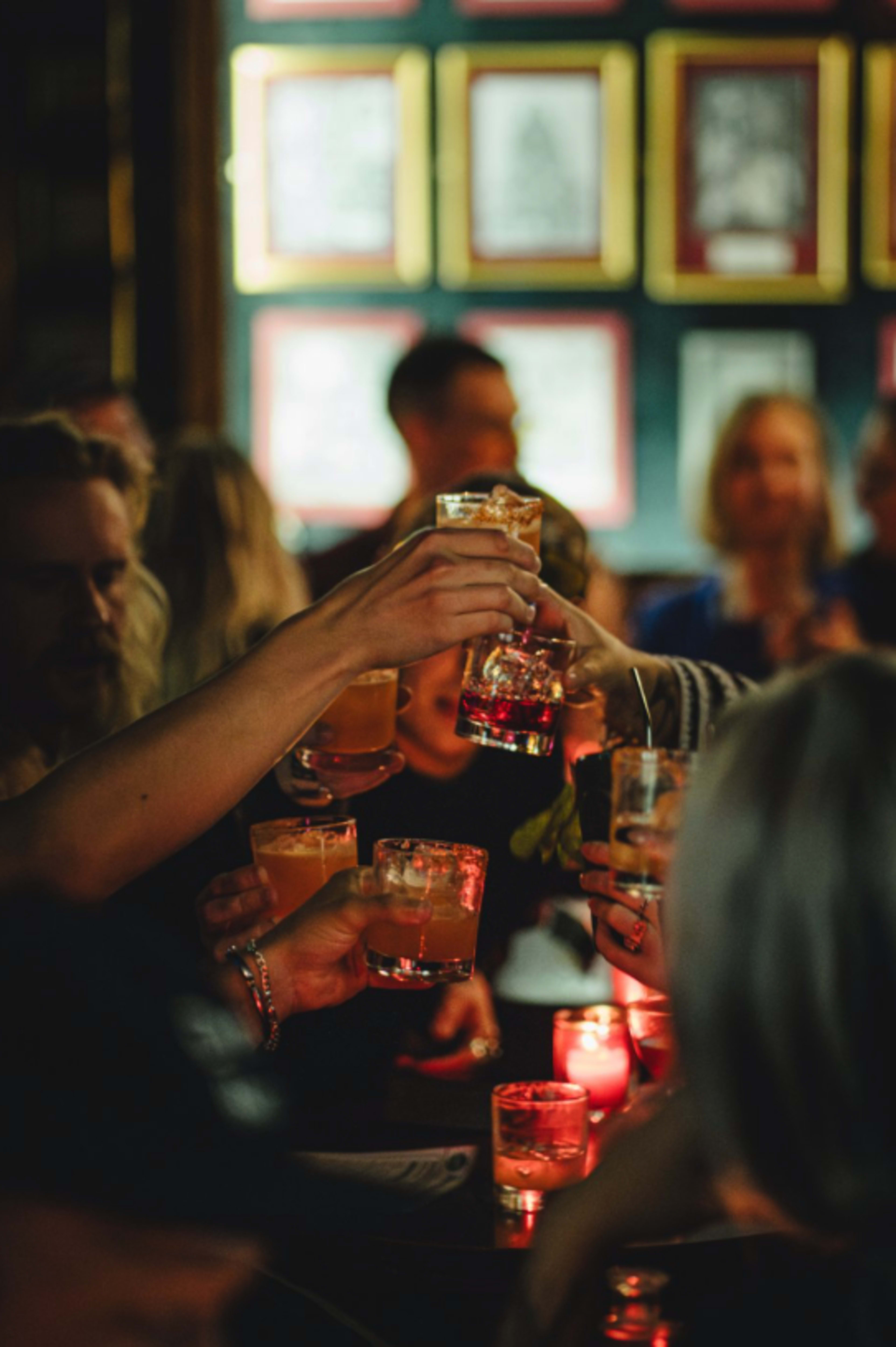 A group of people raises glasses of cocktails in a dimly lit bar, surrounded by framed pictures on the walls.