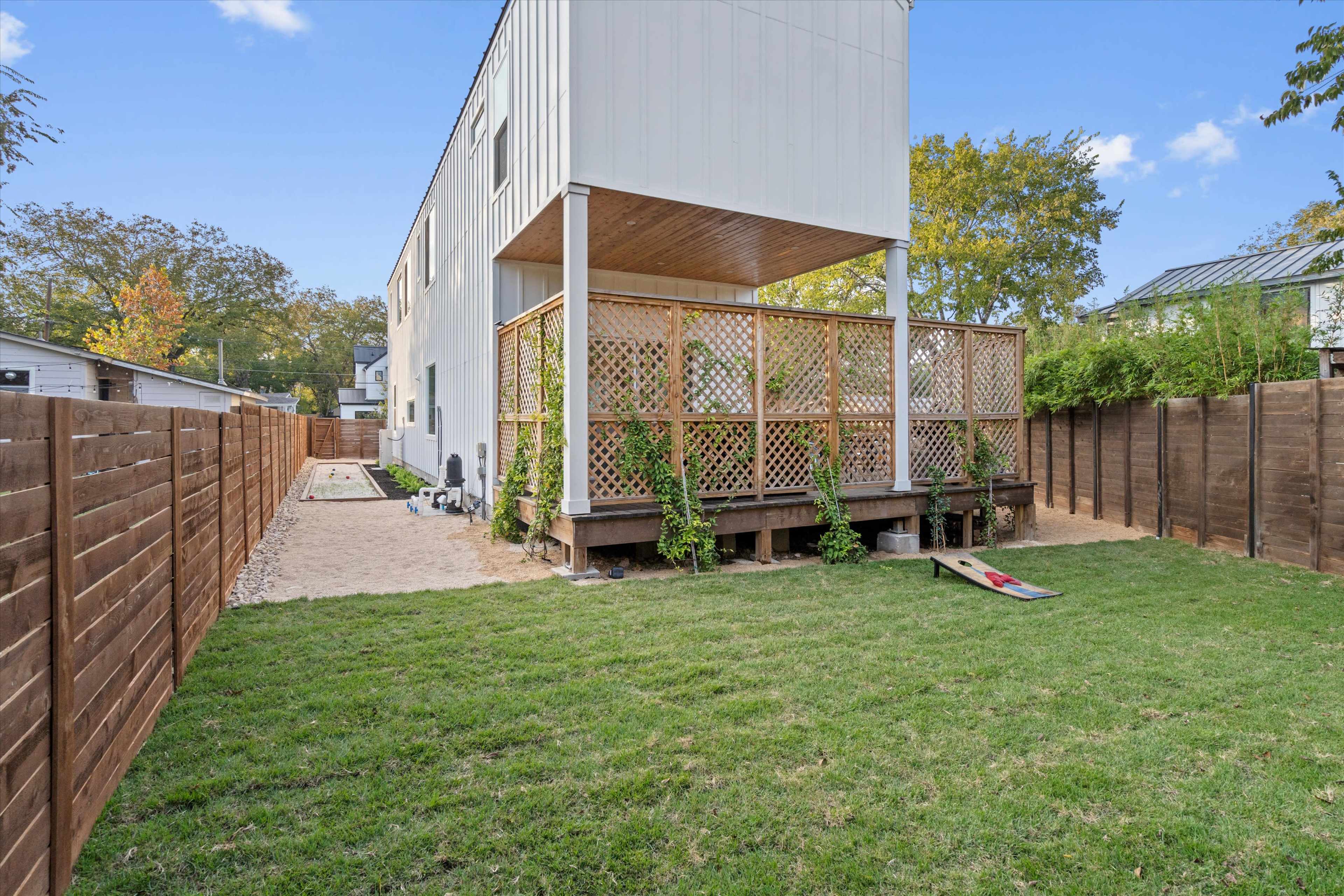 The image shows a backyard with a grassy area, a wooden deck with lattice work, and a pathway leading to the side of a two-story building, surrounded by wooden fences.