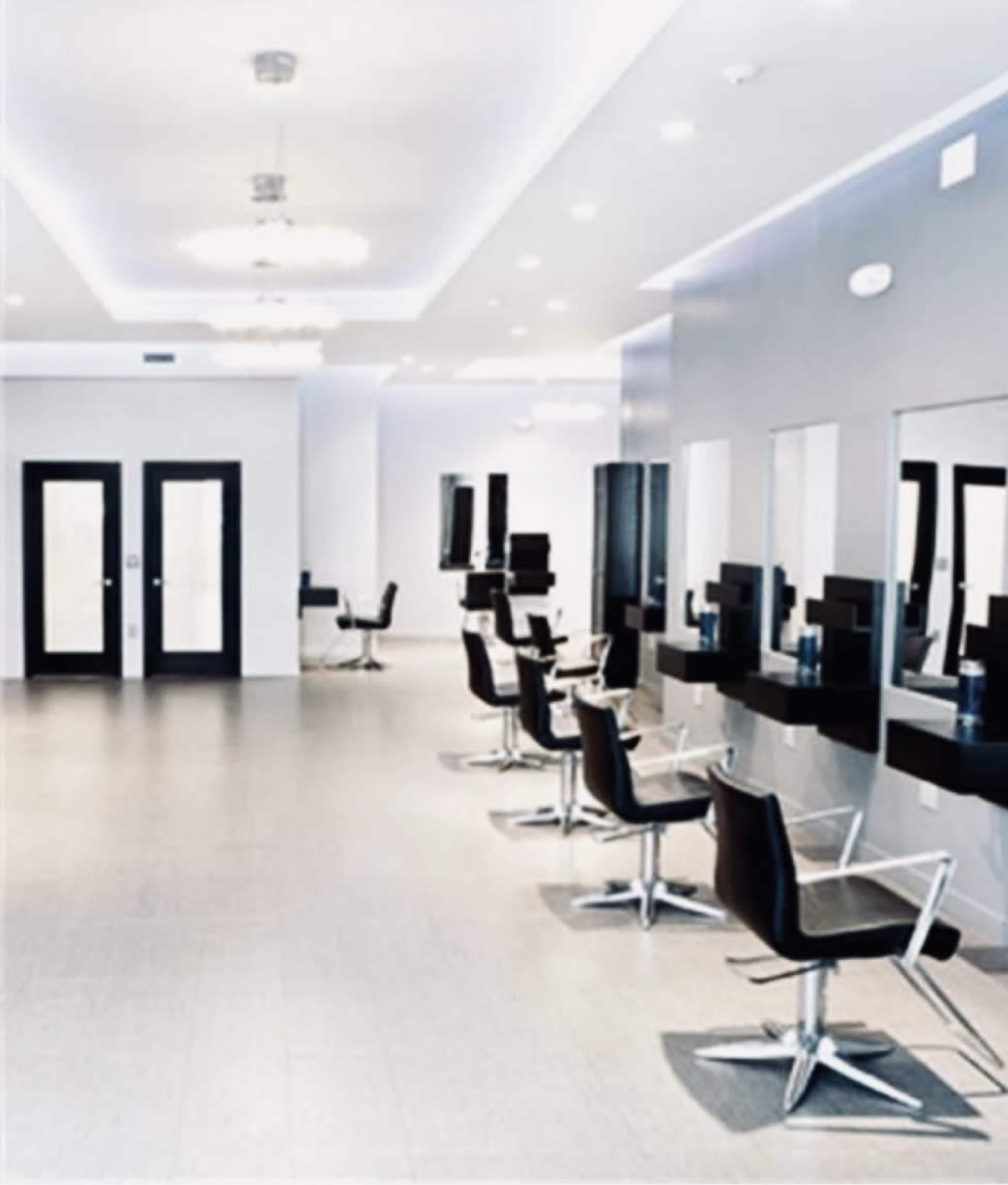 A modern beauty salon features several black salon chairs arranged in front of mirrors along the wall, with sleek lighting and minimalist decor.