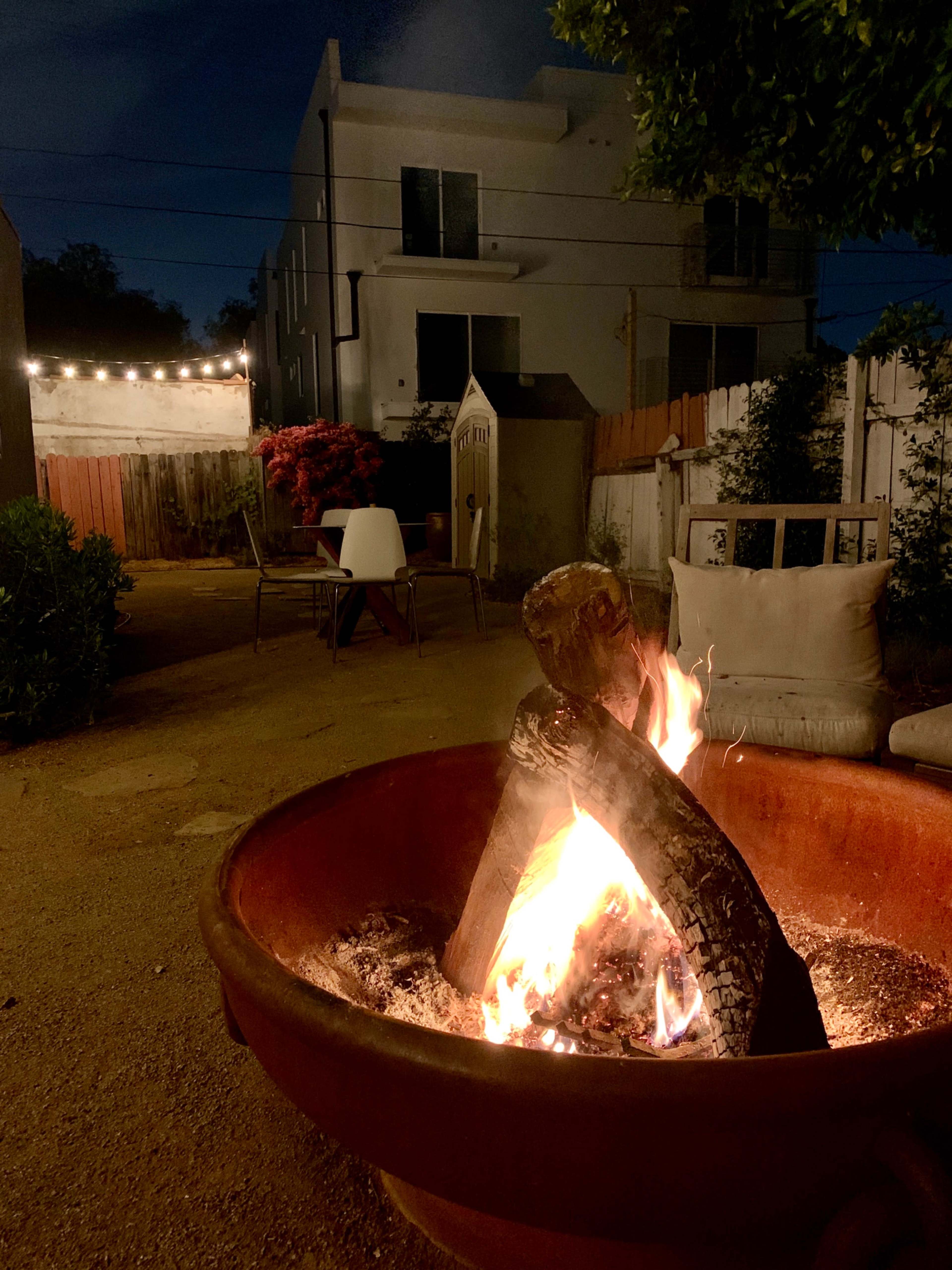 A fire burns in a metal pit in a backyard, with chairs and a small table visible in the background under string lights.
