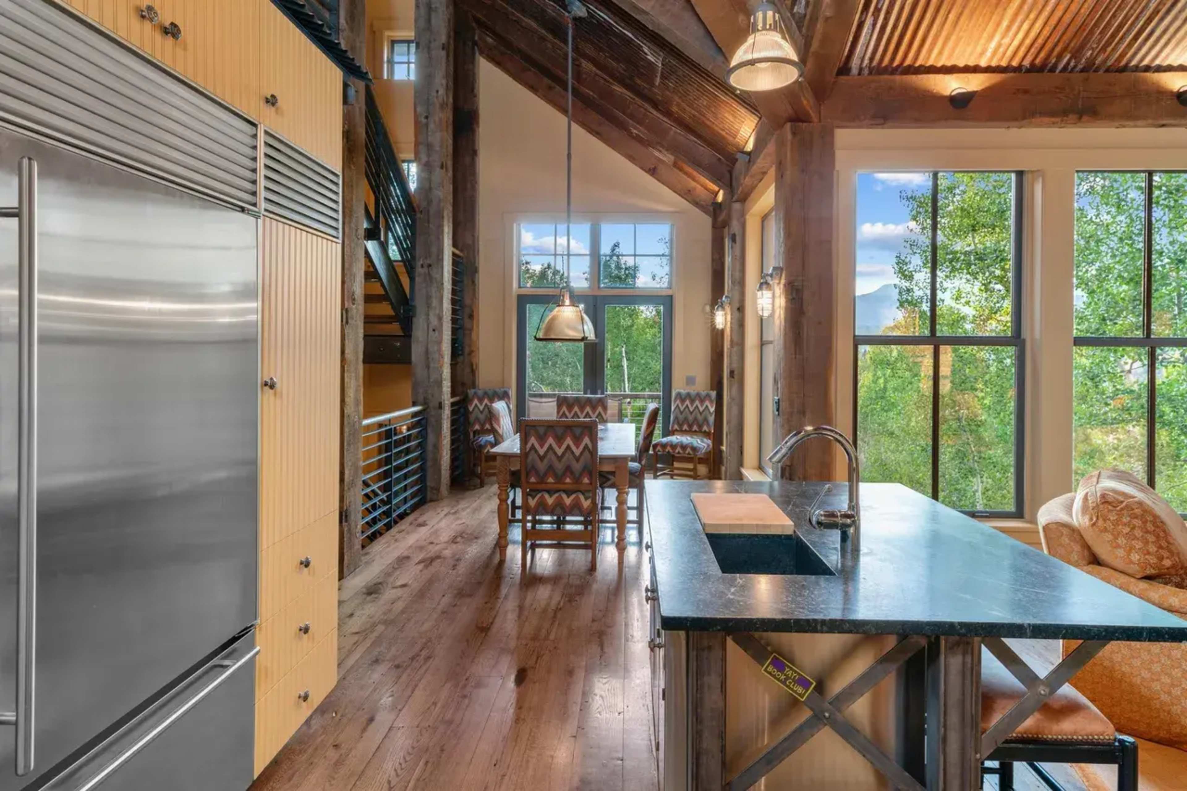 The image shows a modern kitchen with a large island, stainless steel appliances, and large windows overlooking a green landscape.