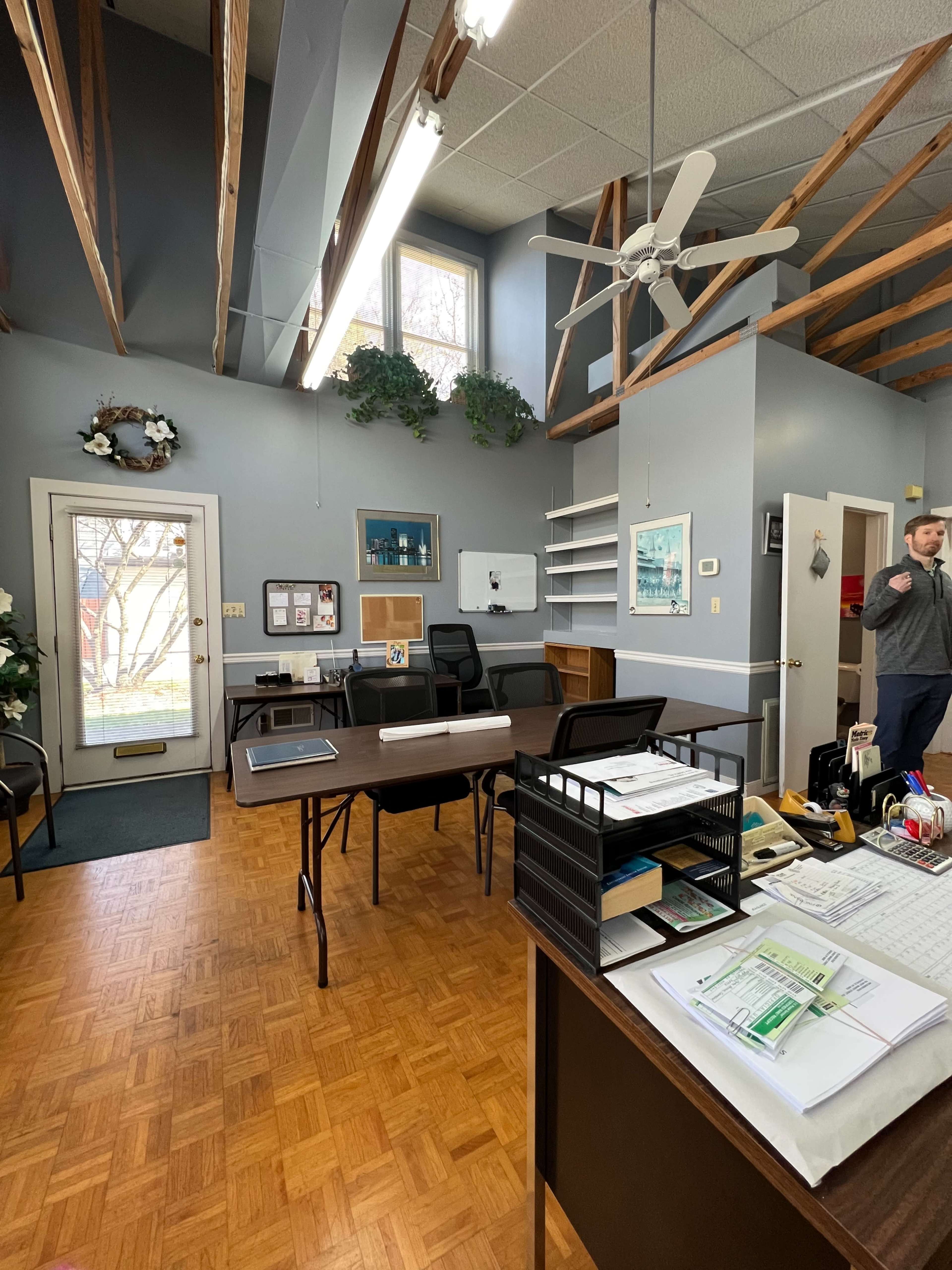 An office space with two tables, chairs, and a door leading to an outdoor area, featuring large windows and wooden beams on the ceiling.